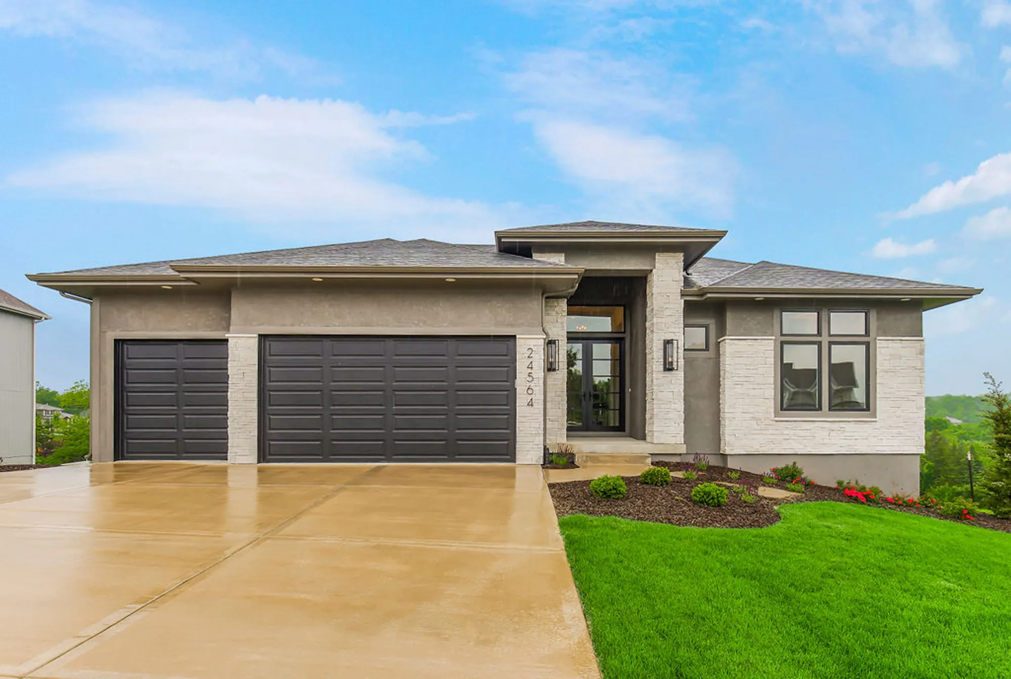 Front view of a modern single-story house with a two-car dark gray garage, stone columns, and a concrete driveway, lawn and sky above.