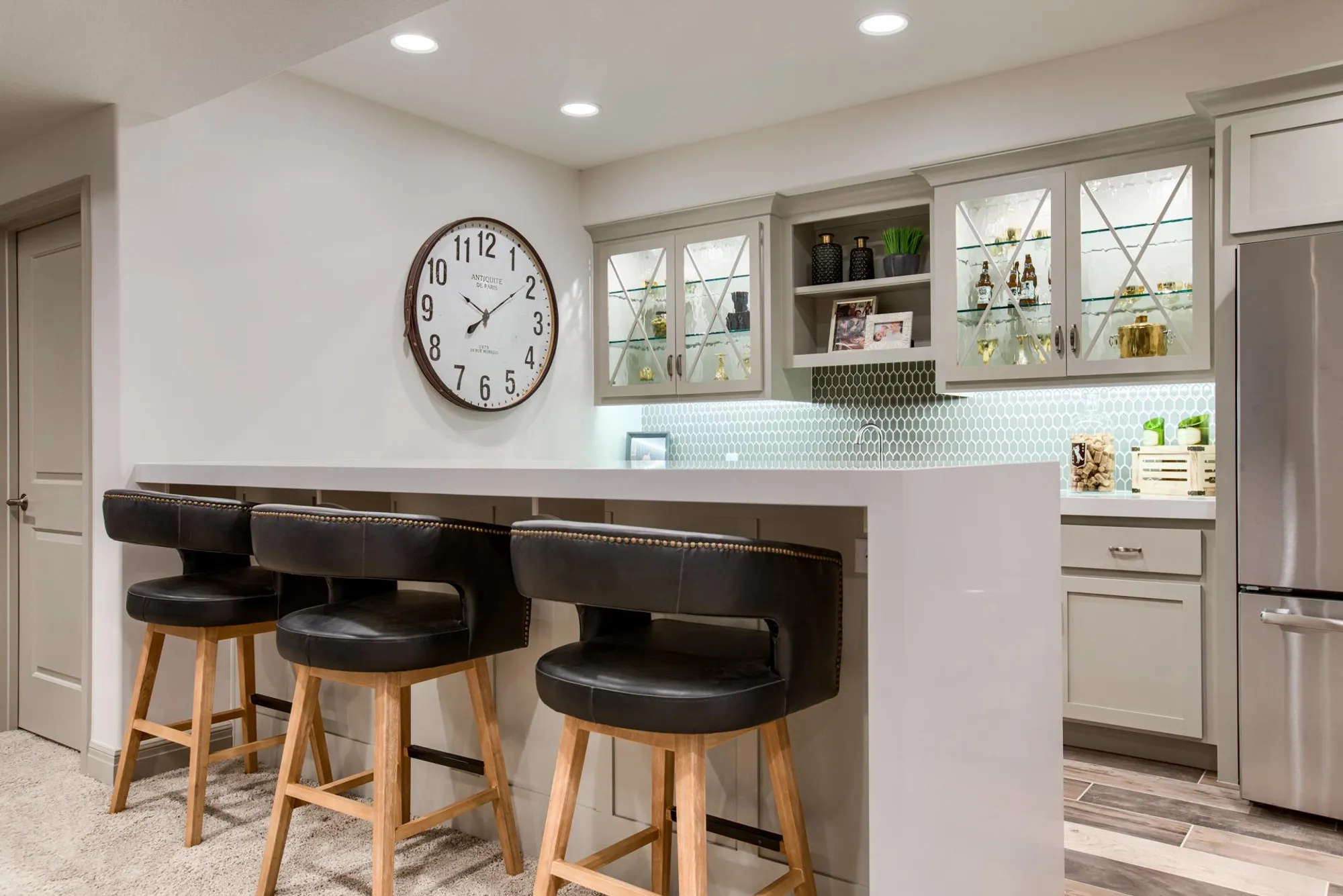 Modern kitchen with gray cabinets, glass-front uppers, a large wall clock, and a white island with black leather bar stools on wooden legs.