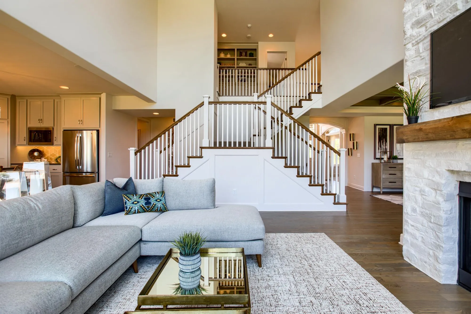 Open-concept living room with a gray sectional, glass coffee table, and a grand white staircase centered in the space.