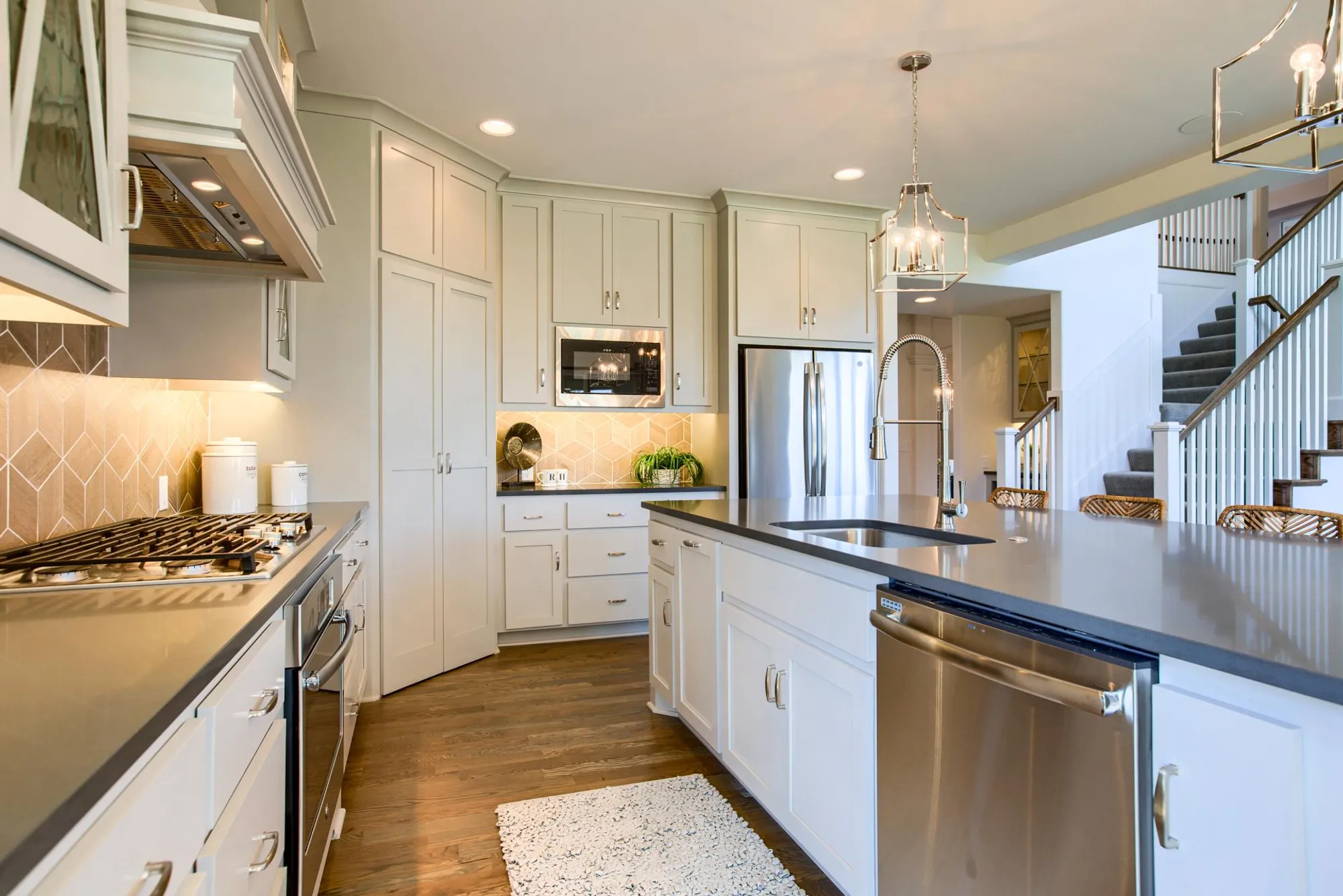 Bright modern kitchen with white cabinets, stainless steel appliances, and a large island with a sink; staircase visible in the background.
