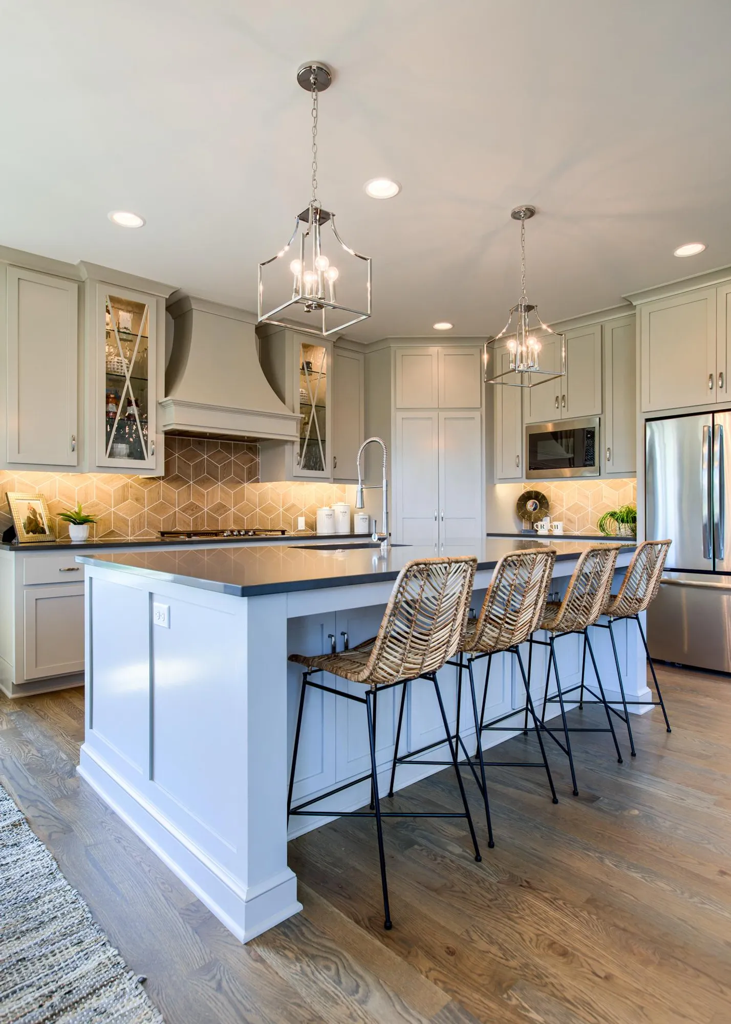 Bright, modern kitchen with an island, light gray cabinets, woven bar stools, and two geometric pendant lights above.