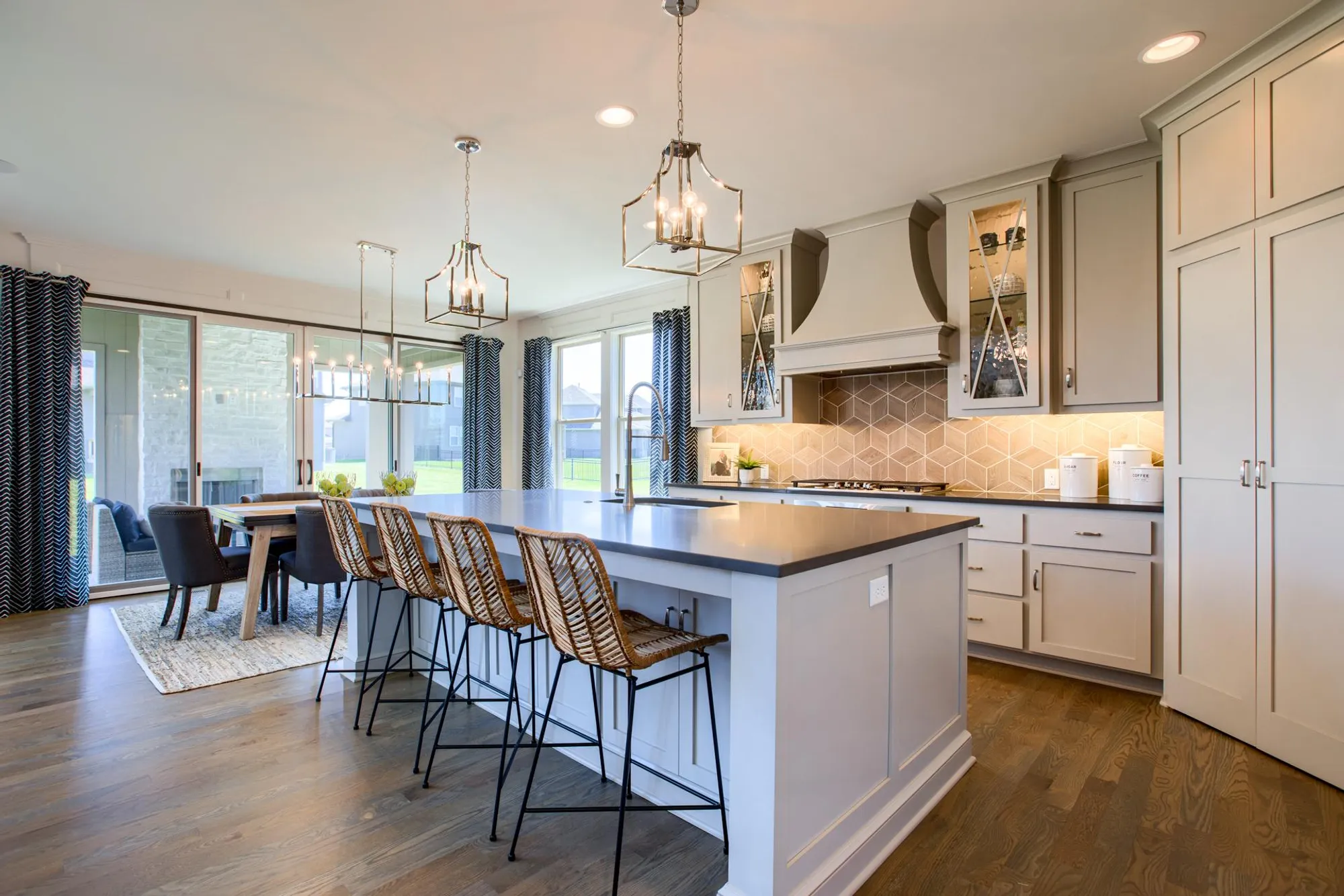 Bright open kitchen with a large island, pendant chandeliers, and gray cabinetry along a hex tile backsplash