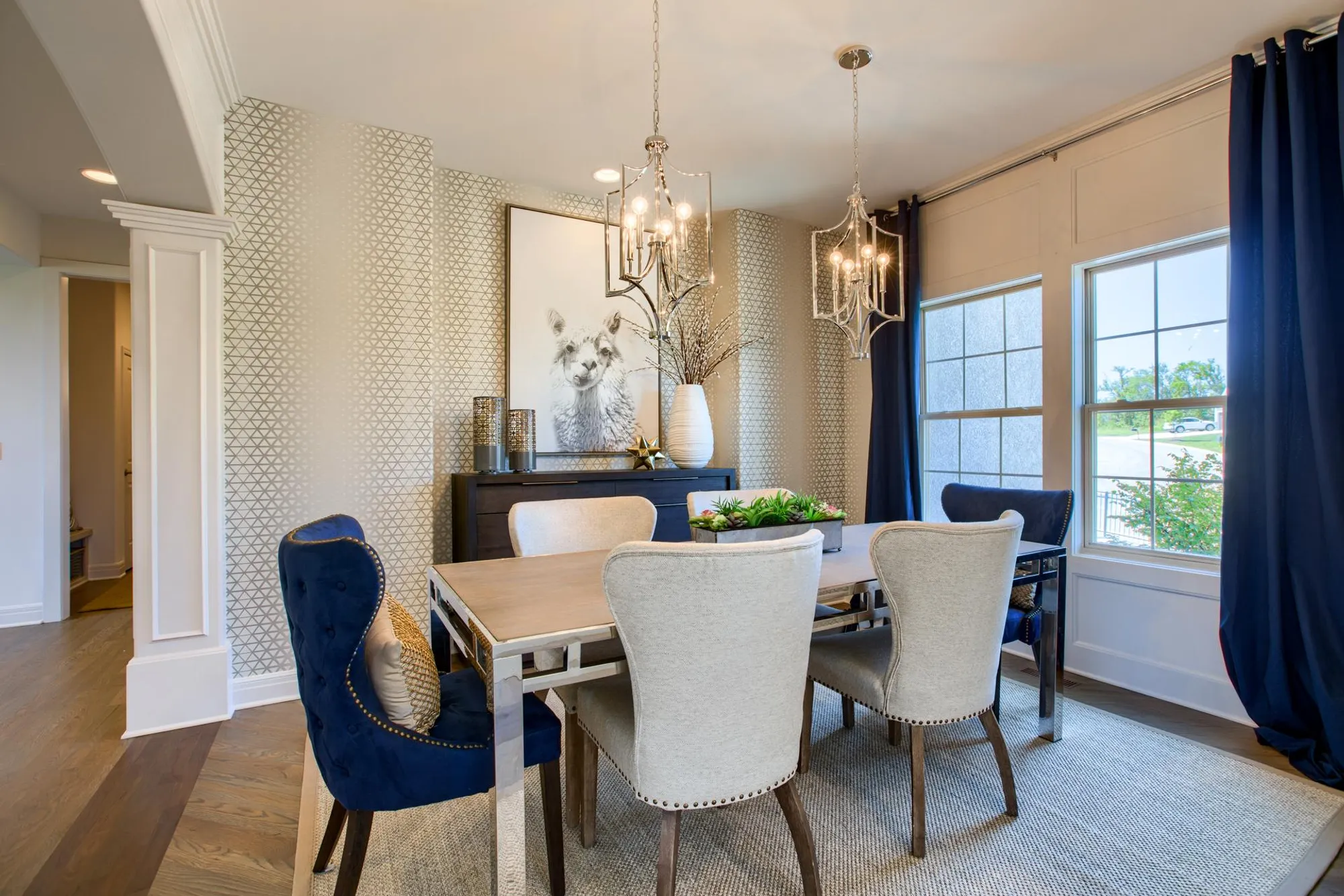 Dining room with a dark sideboard, llama artwork, crystal chandeliers, and a light wood table surrounded by beige and navy chairs; patterned wallpaper and large windows with blue curtains.
