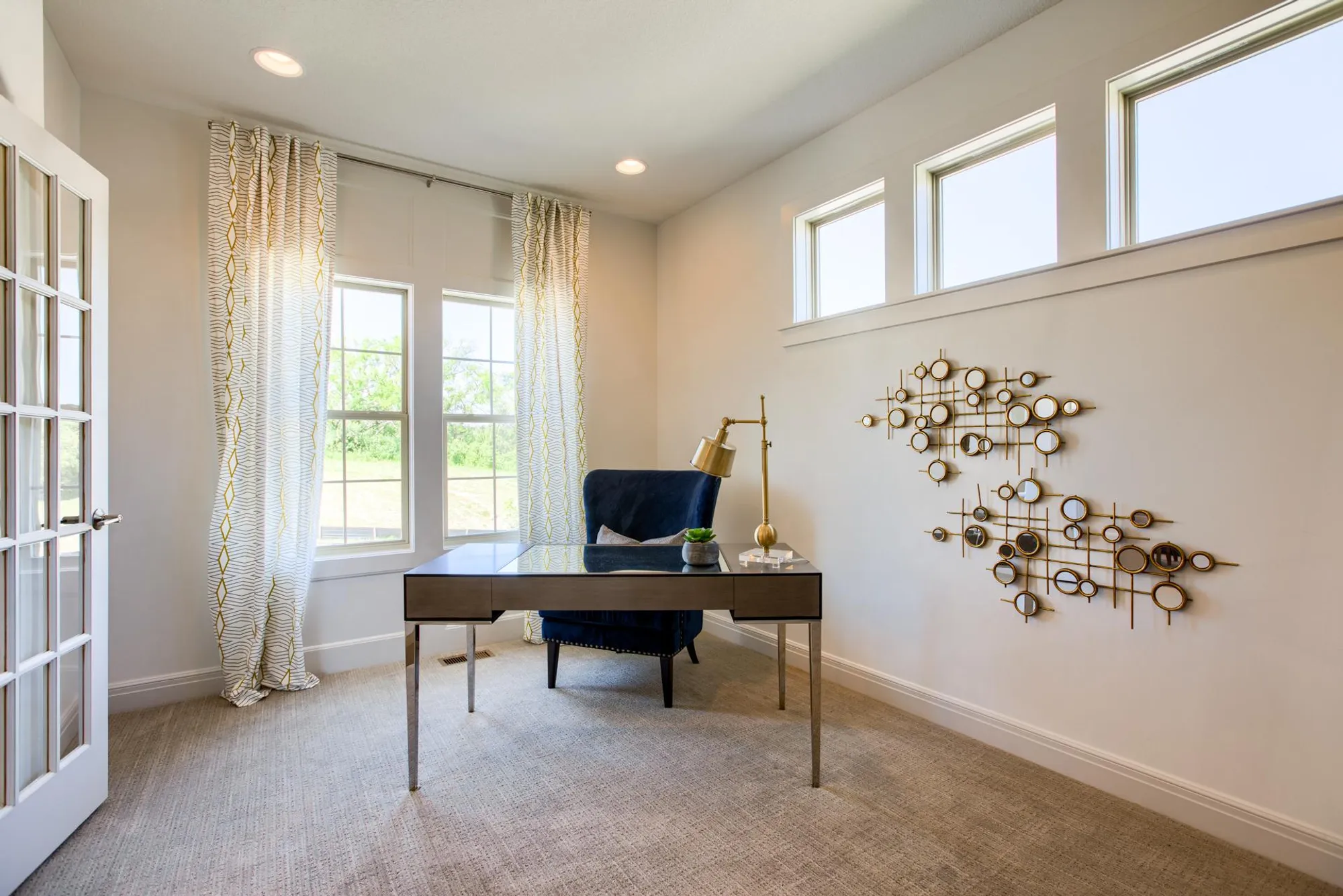 Bright home office with a glass desk, blue upholstered chair, gold desk lamp, and a geometric circular wall sculpture; large windows provide natural light.
