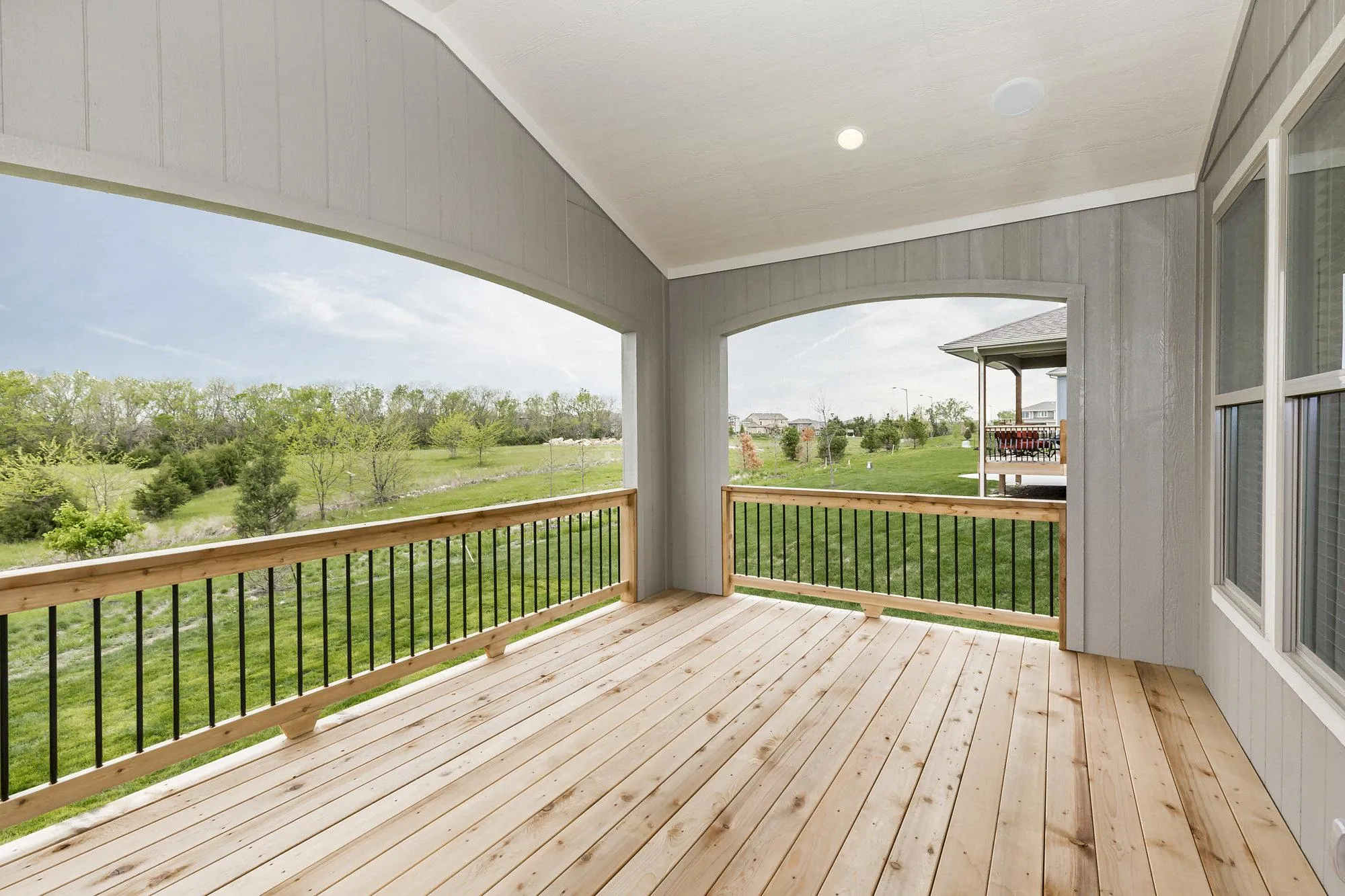 Covered wooden balcony with railings overlooking a green yard and trees, exterior siding, and a neighboring house in the distance; bright outdoor lighting overhead