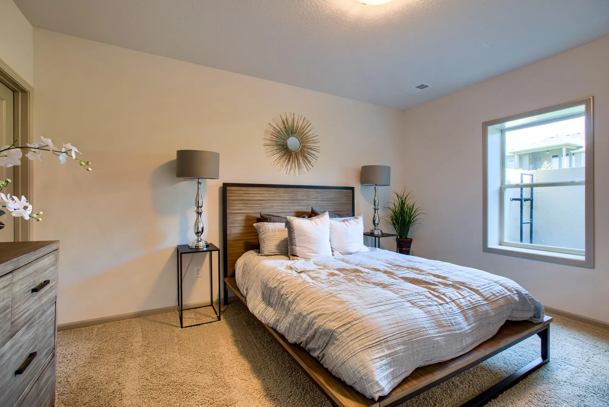 Modern bedroom with a low wooden platform bed, two metal nightstands and silver lamps, a sunburst mirror above the headboard, and a large window to the right.