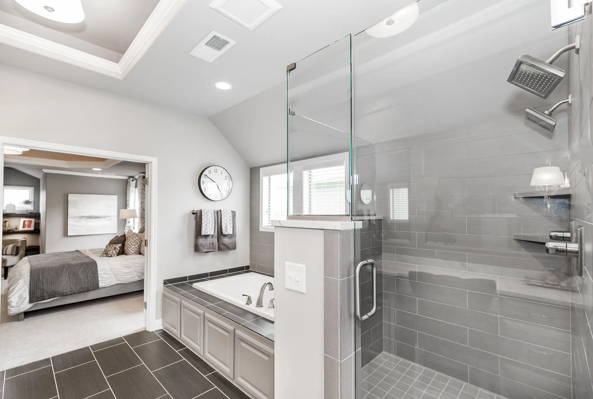 Modern bathroom with a glass shower enclosure, gray tile, and chrome fixtures; bedroom visible through the doorway.