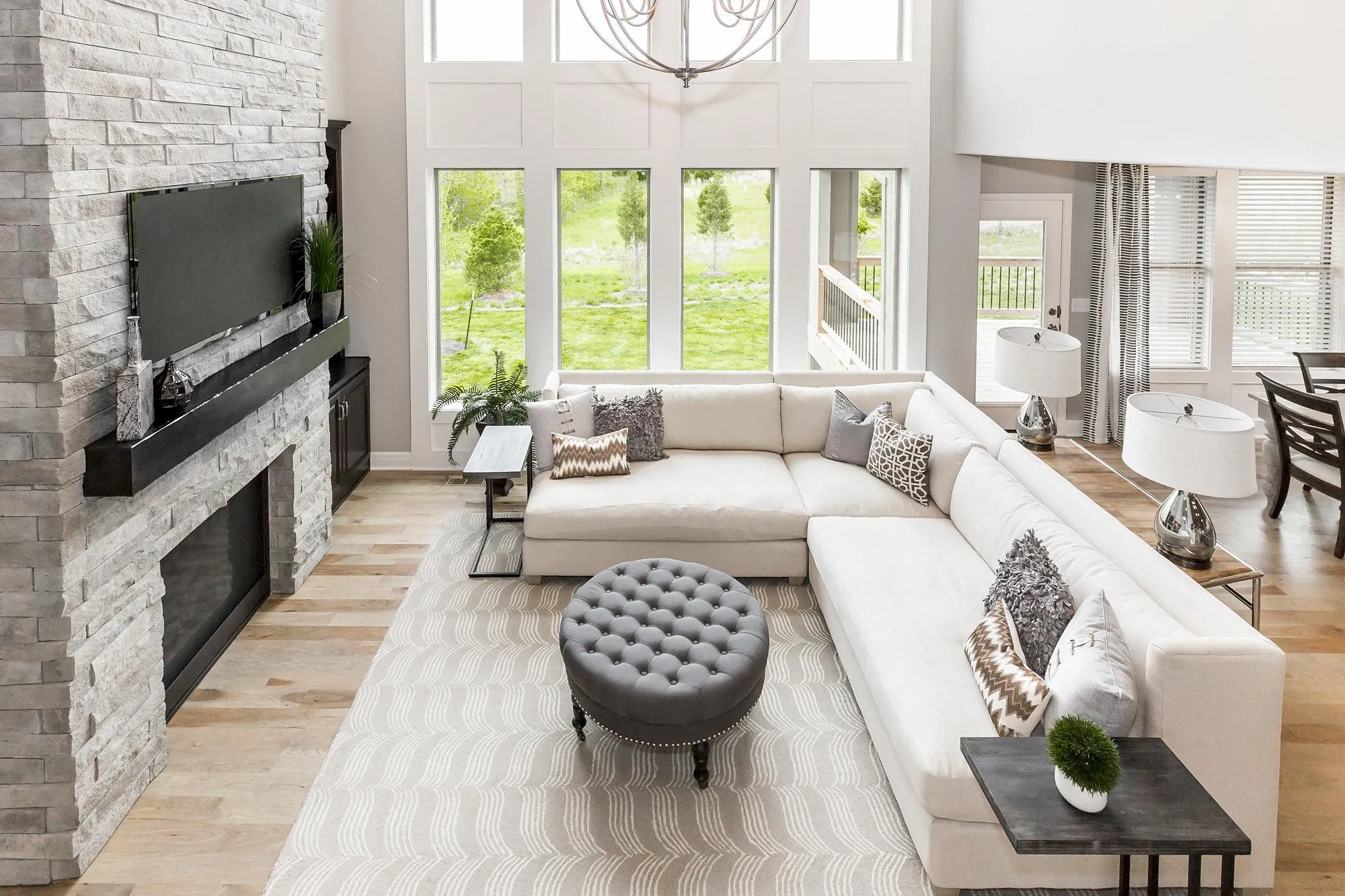 Bright living room with a white sectional sofa, gray patterned rug, and tufted round ottoman facing a stone fireplace and wall-mounted TV; large windows let in natural light and overlook a green outdoor space.