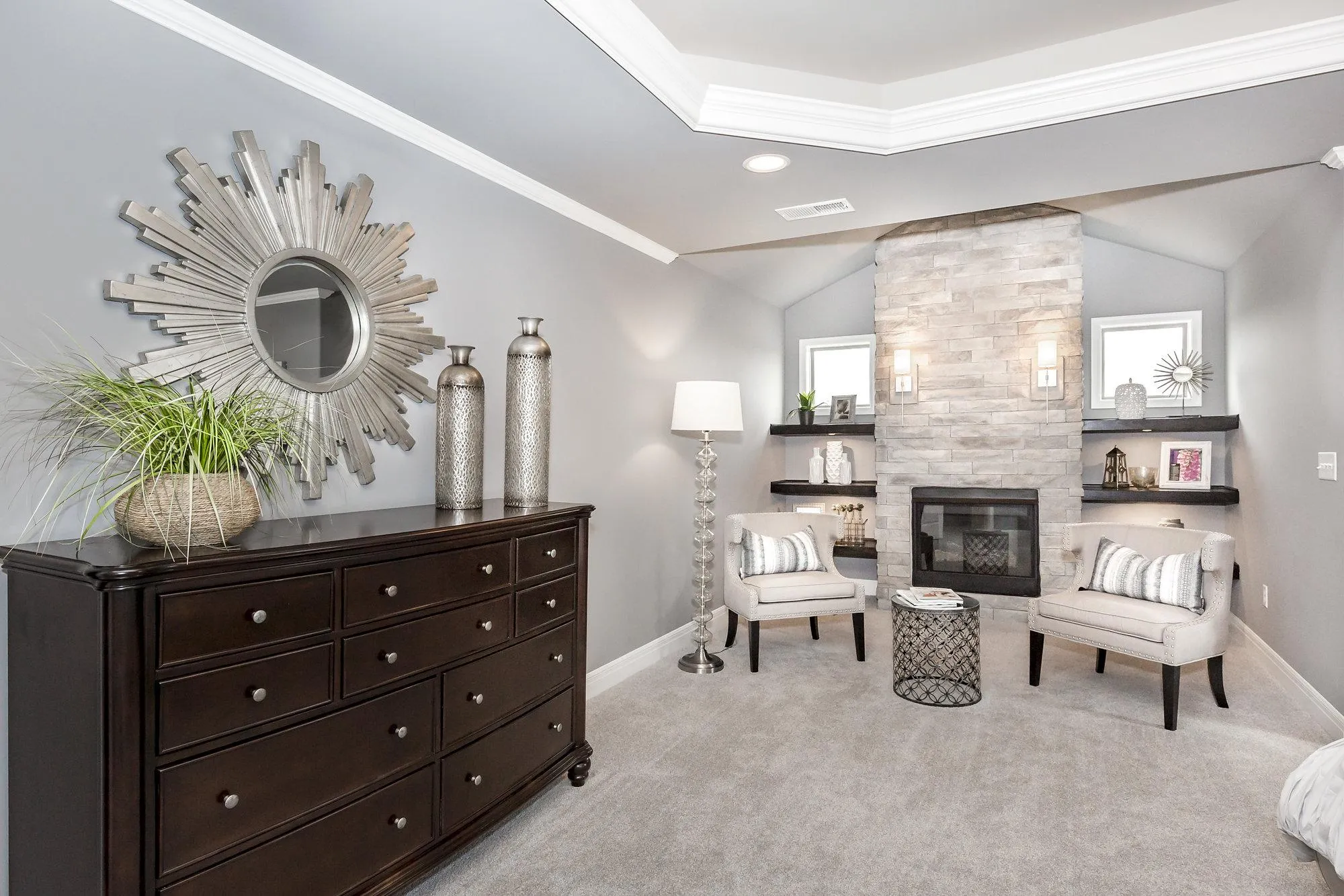 Living room with a dark wooden dresser, a round sunburst mirror, greenery, and two tall metallic vases against a gray wall; a stone fireplace and shelves to the right create a modern, elegant focal point.
