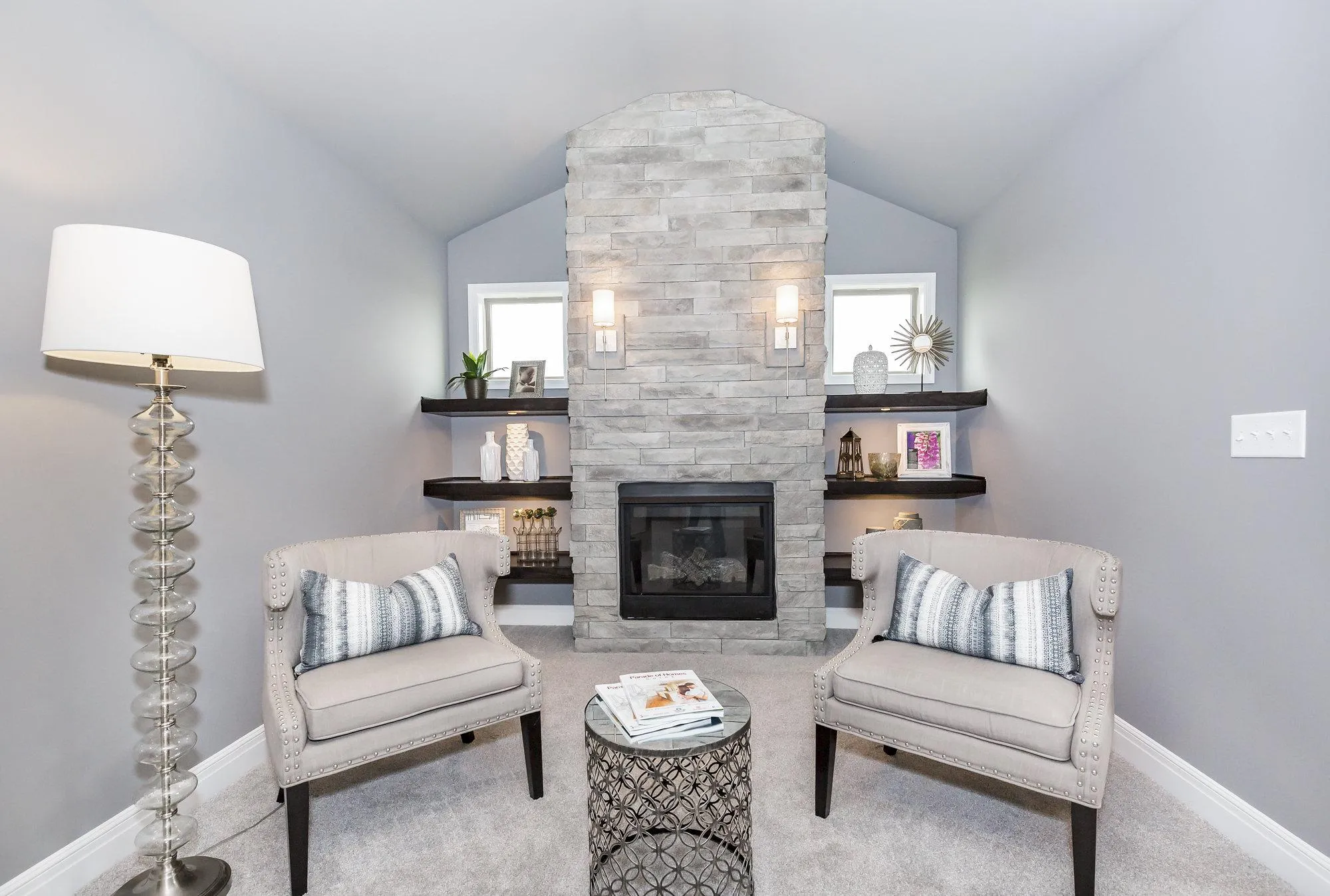 Cozy living room seating area with two upholstered armchairs facing a stone fireplace and a round metal side table between them on a gray carpeted floor.