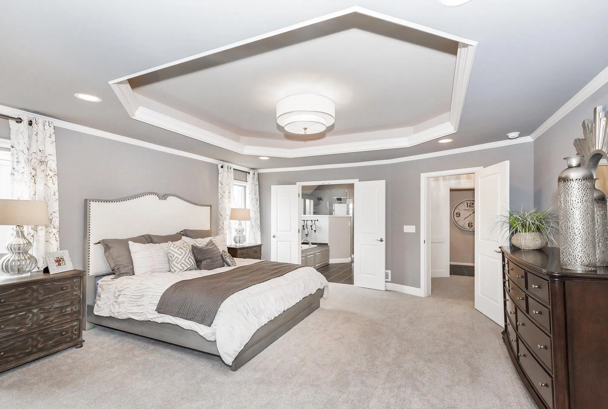 Modern gray bedroom featuring a large bed, white bedding, and two dark wood dressers under a tray ceiling with a central light fixture.