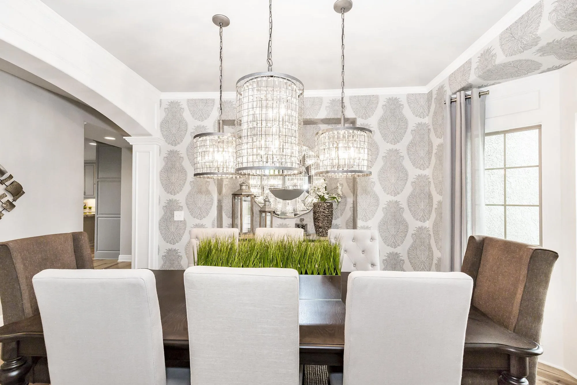 Dining room with three crystal chandeliers over a dark wood table and gray upholstered chairs, gray patterned wallpaper in the background.