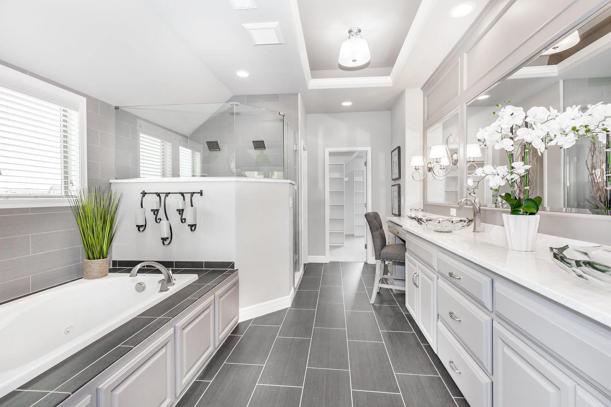 Bright modern bathroom with double vanity, large mirror, glass shower, tub, gray tiled floor, and a potted orchid plant.