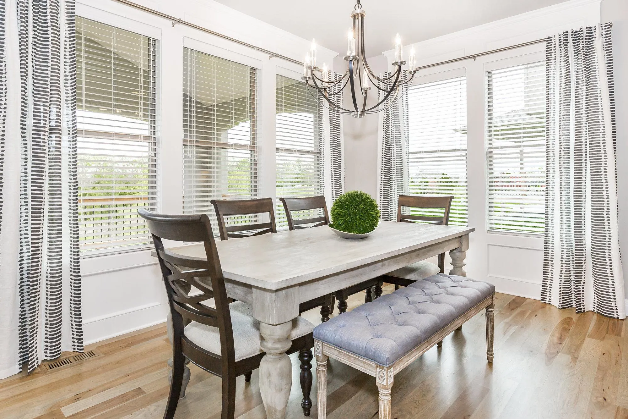 Bright dining nook with a rustic wood table, dark chairs, a cushioned bench, striped curtains, and a black chandelier overhead.