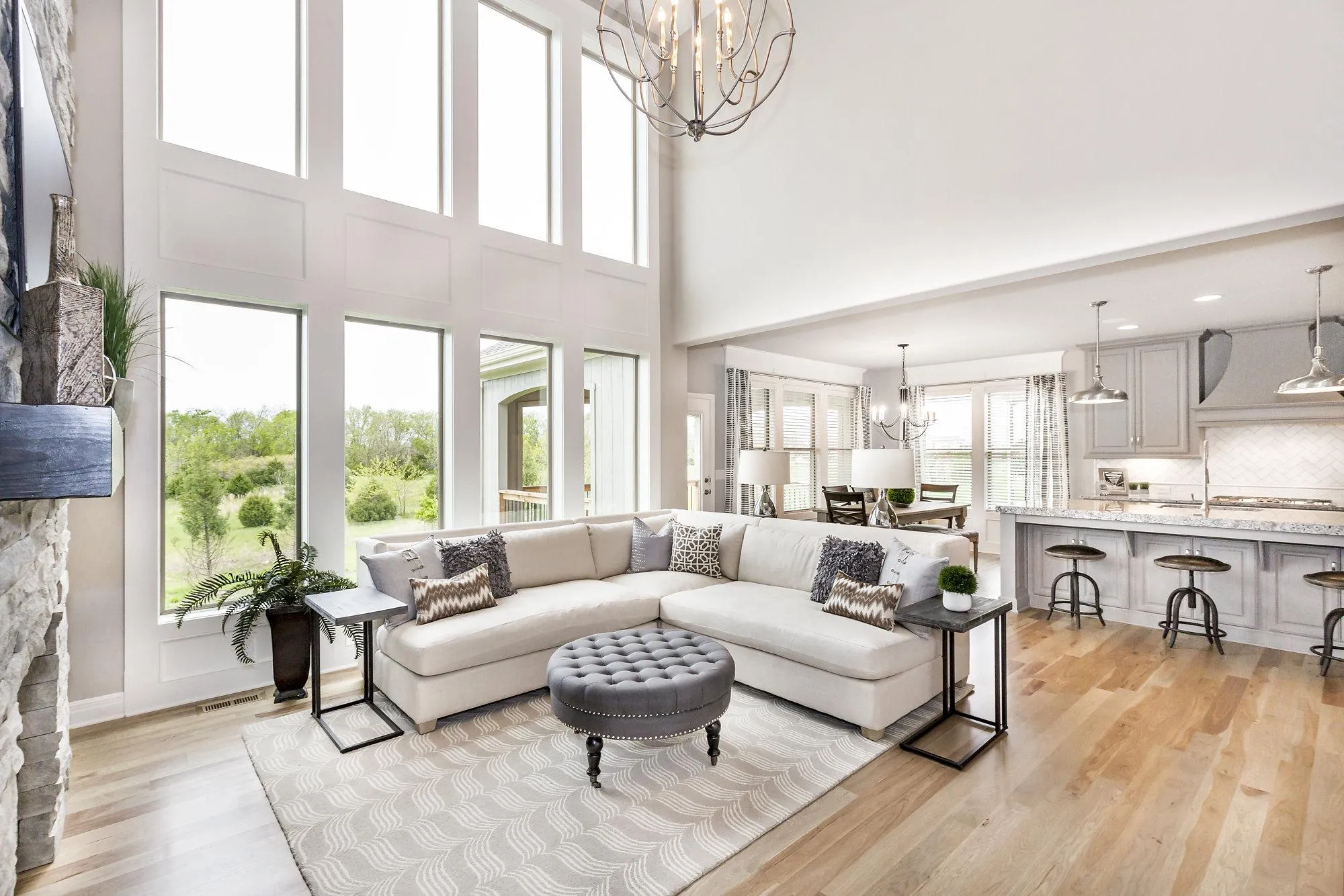 Bright open living room with oversized windows, a light-gray sectional sofa, tufted gray ottoman, and a modern kitchen in the background.