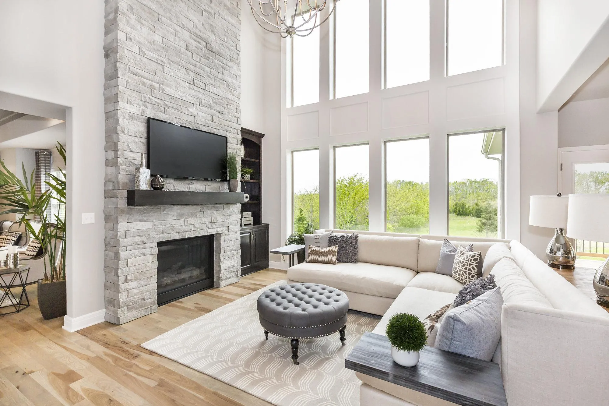 Bright living room with a stacked stone fireplace, mounted TV, and large windows behind a light-colored sectional.