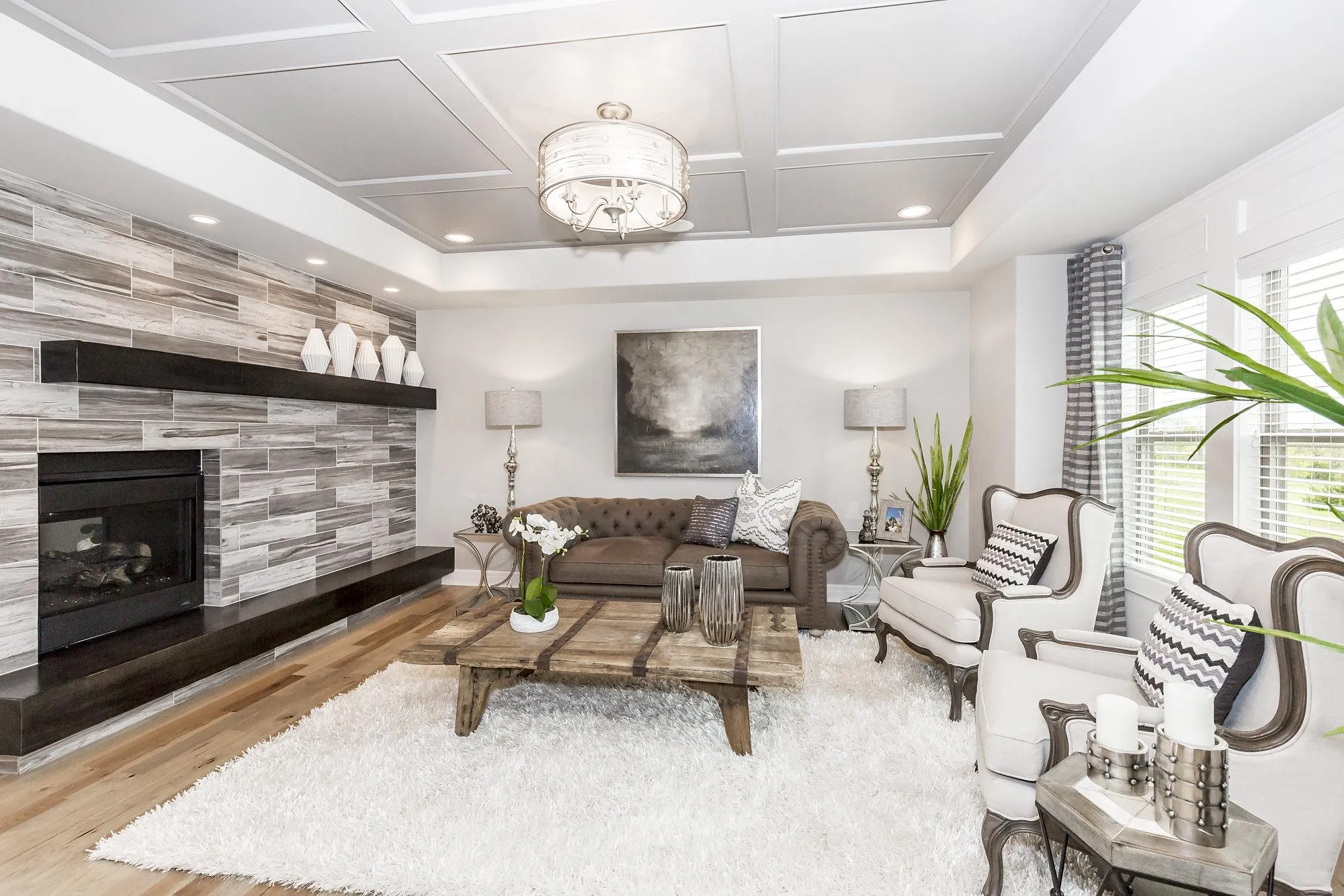 Bright living room with a gray-tiled fireplace wall, white walls, and a fluffy white rug; brown tufted sofa and white accent chairs nearby.