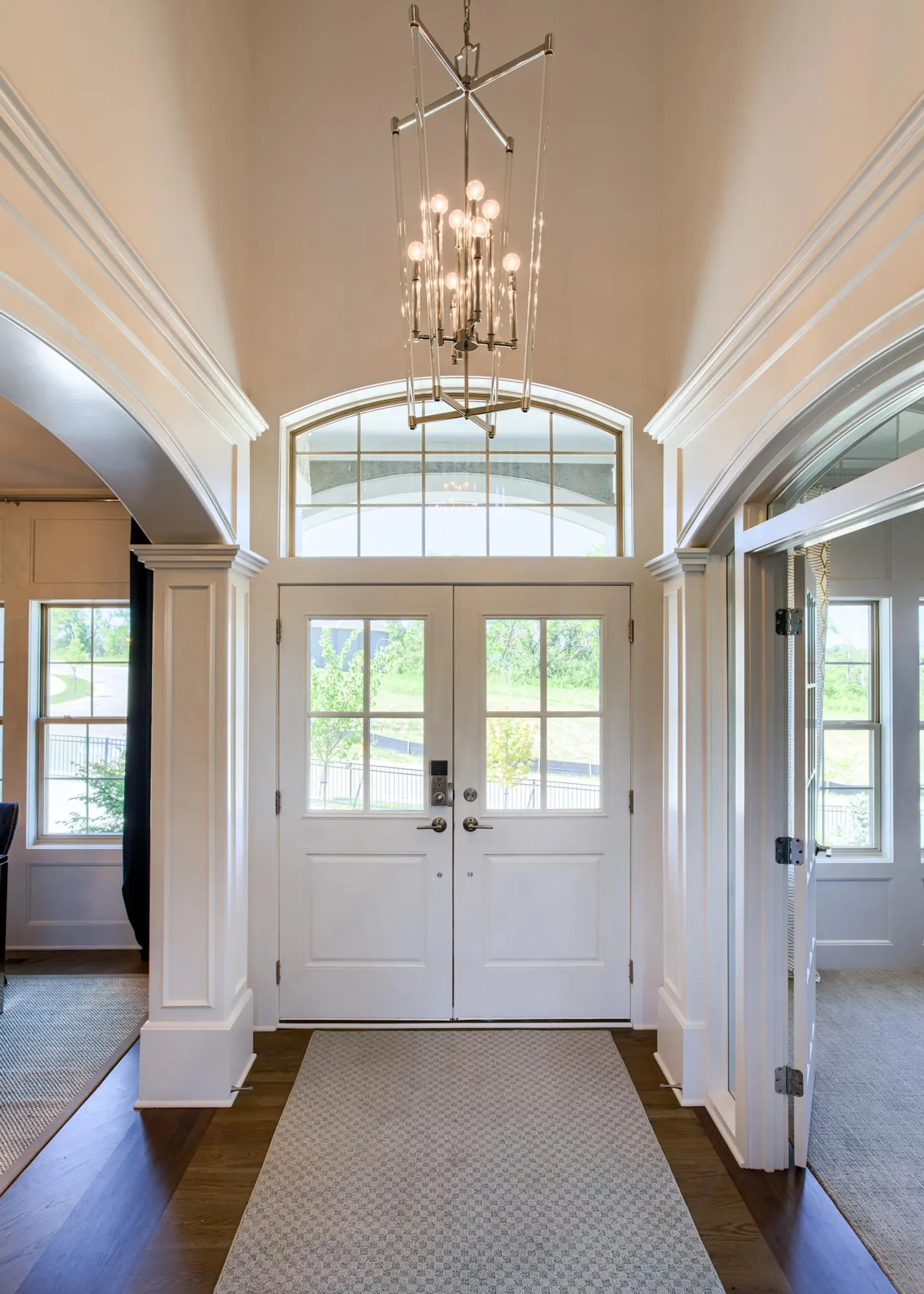 Bright entryway with double white doors, large arched window above, and a modern geometric chandelier hanging from the ceiling.