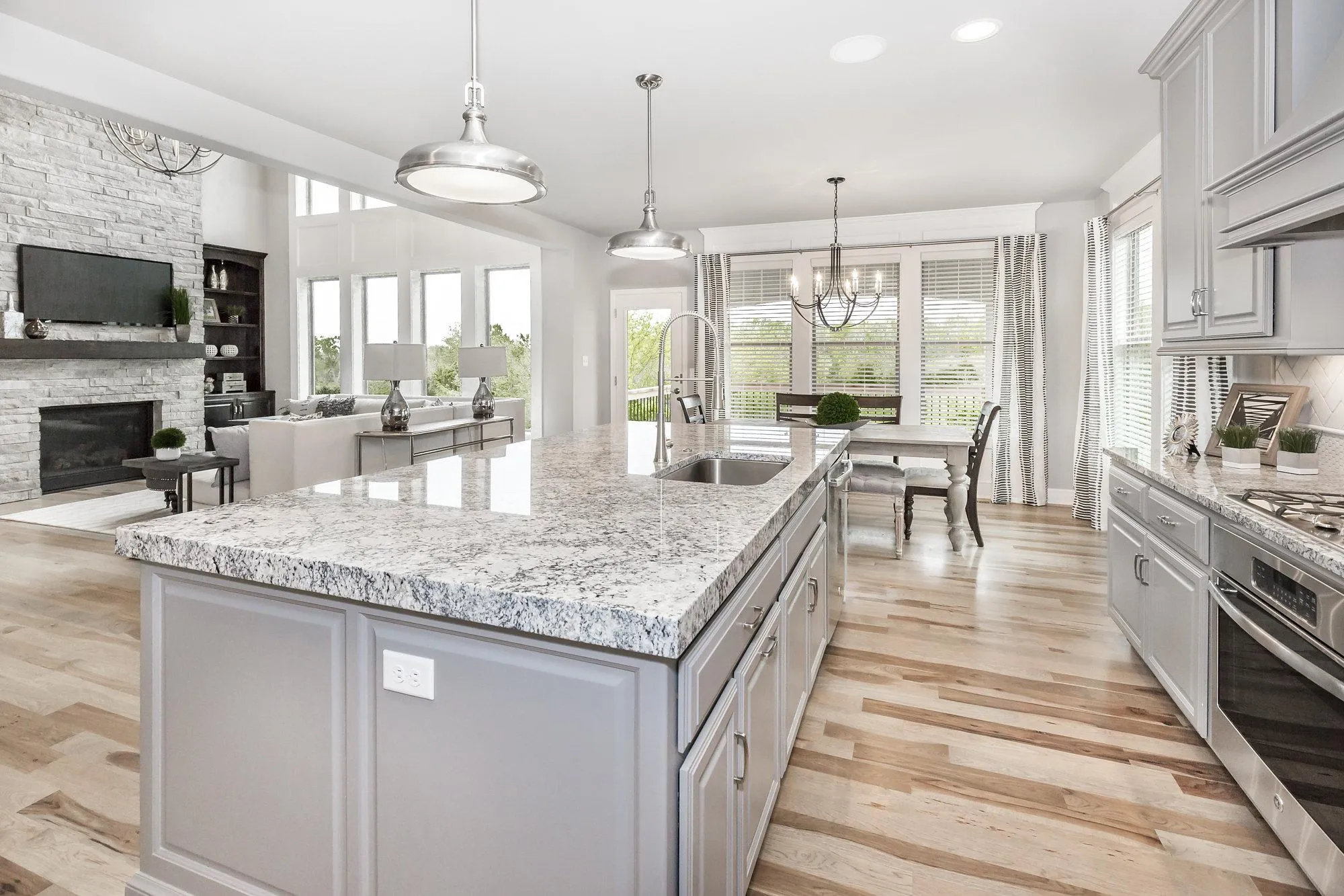 Open-concept kitchen with grey cabinetry, a large granite island, stainless appliances, and a view into the living area with a stone fireplace.