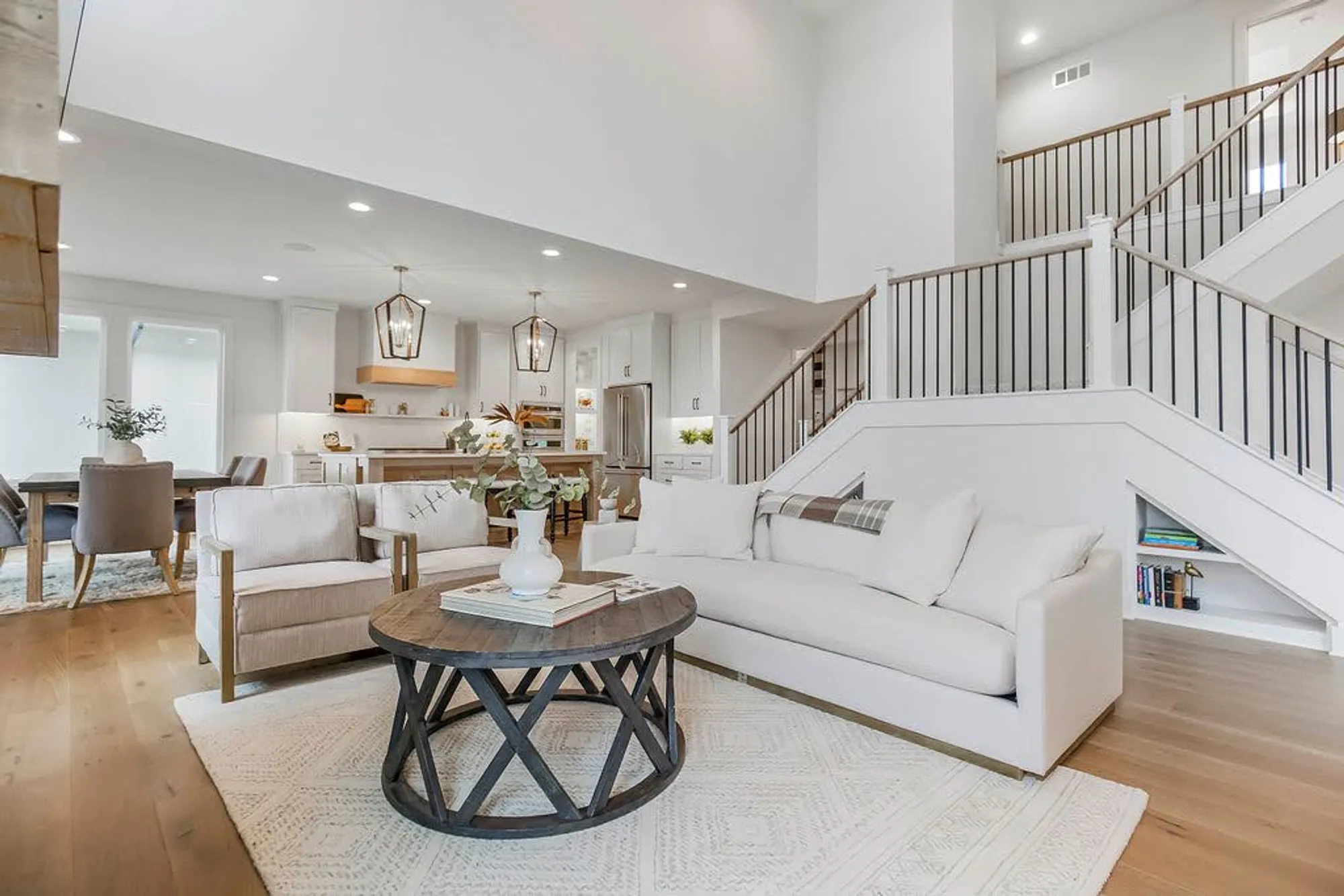 Bright open living space with a white kitchen, beige seating, and a round coffee table; staircase with black railings in the background.