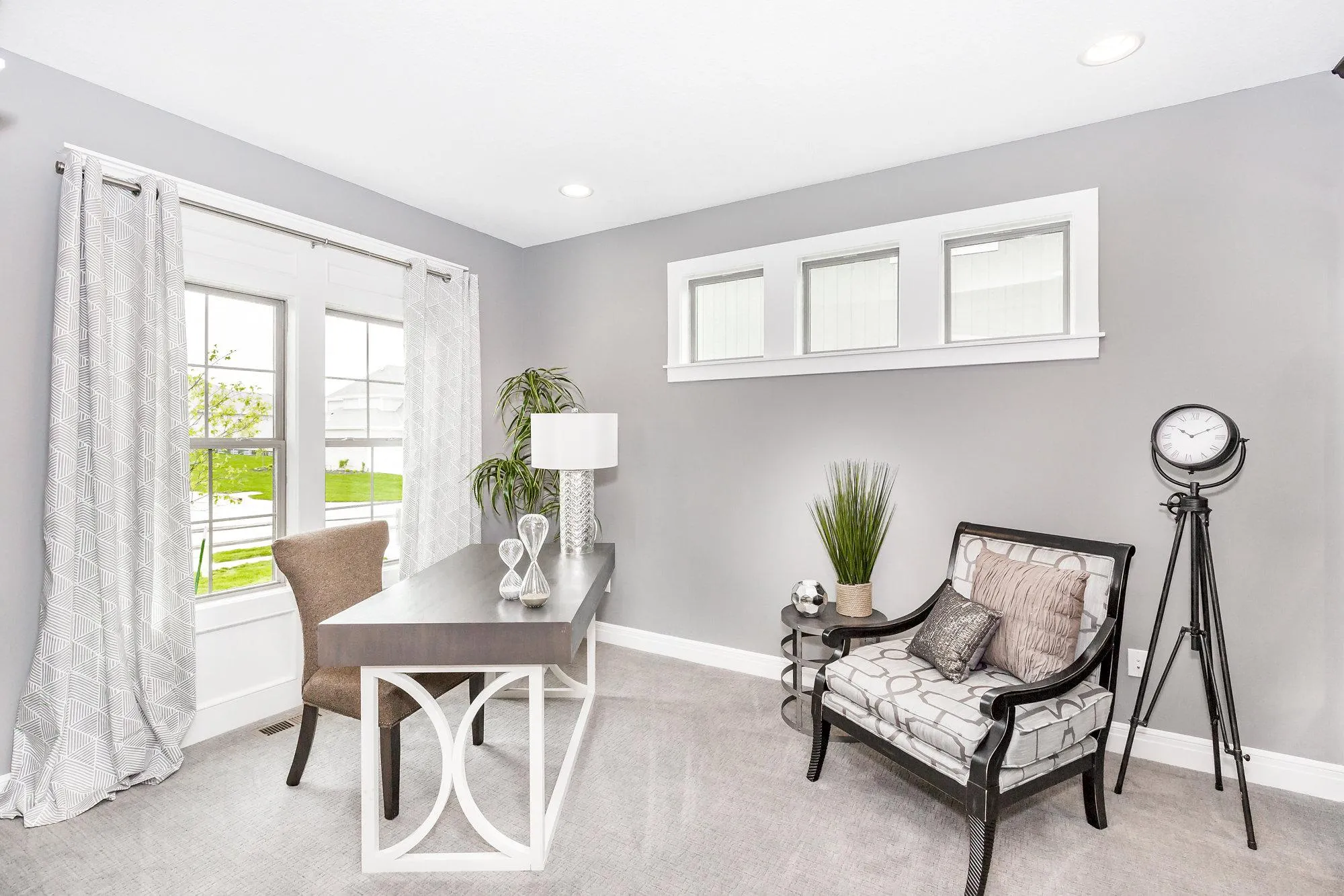 Modern living room corner with gray walls, large window with geometric curtains, a white-framed desk, beige chair, and tall plants.