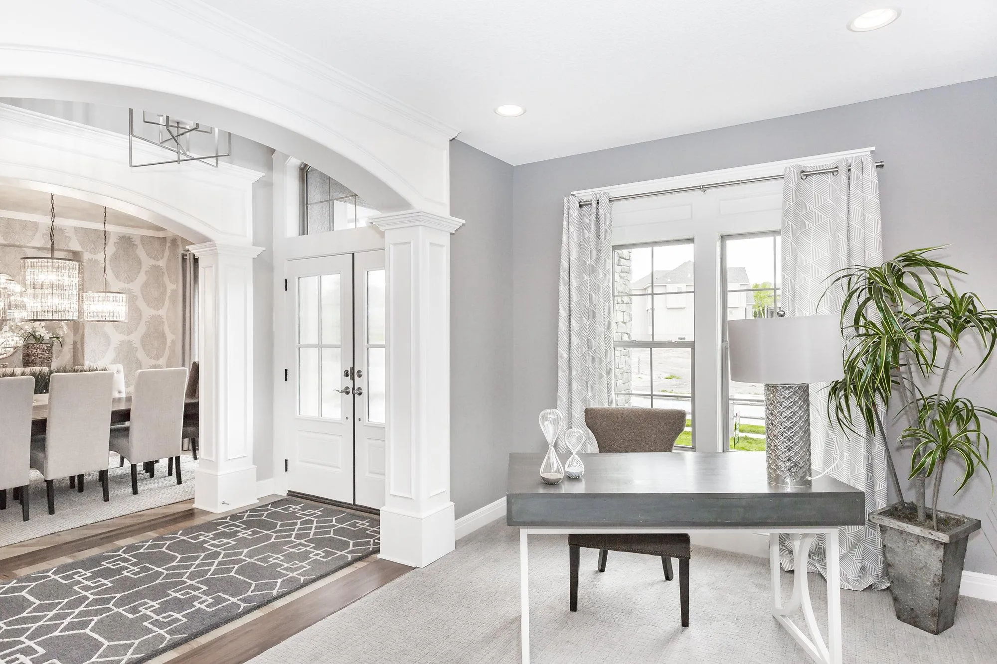 Open-concept dining and living area with white arches, gray walls, and a split-level desk near large windows with patterned curtains