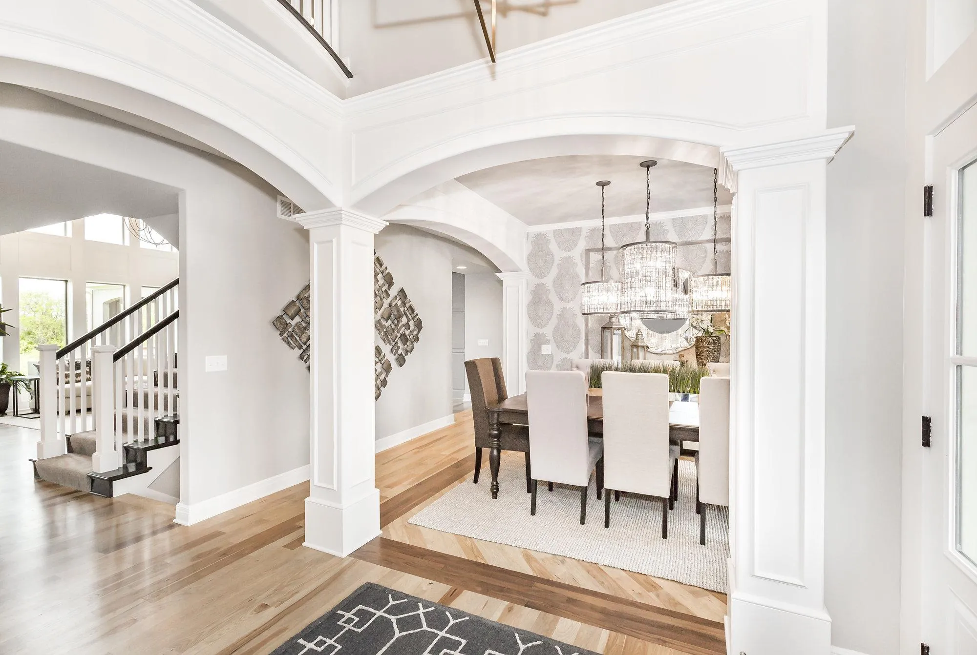 Bright two-story foyer with white arches and columns, view into a dining area with a large crystal chandelier and patterned wallpaper in the background.