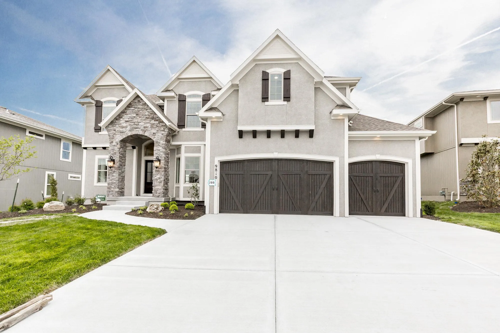 Two-story gray suburban home with stone entry, dark brown garage doors, and manicured lawn under a blue sky.
