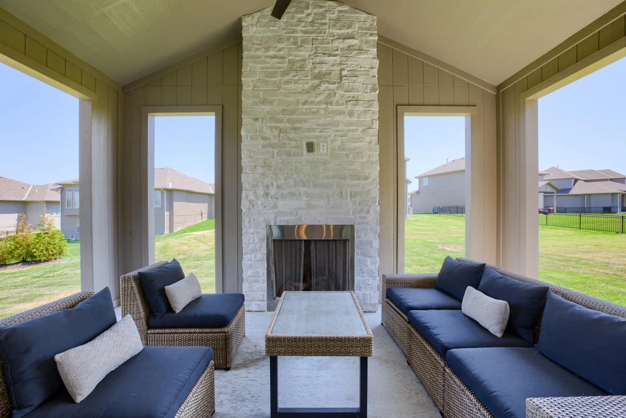 Outdoor covered patio with a central stone fireplace, wicker seating and a glass coffee table, overlooking a green yard and neighboring houses.