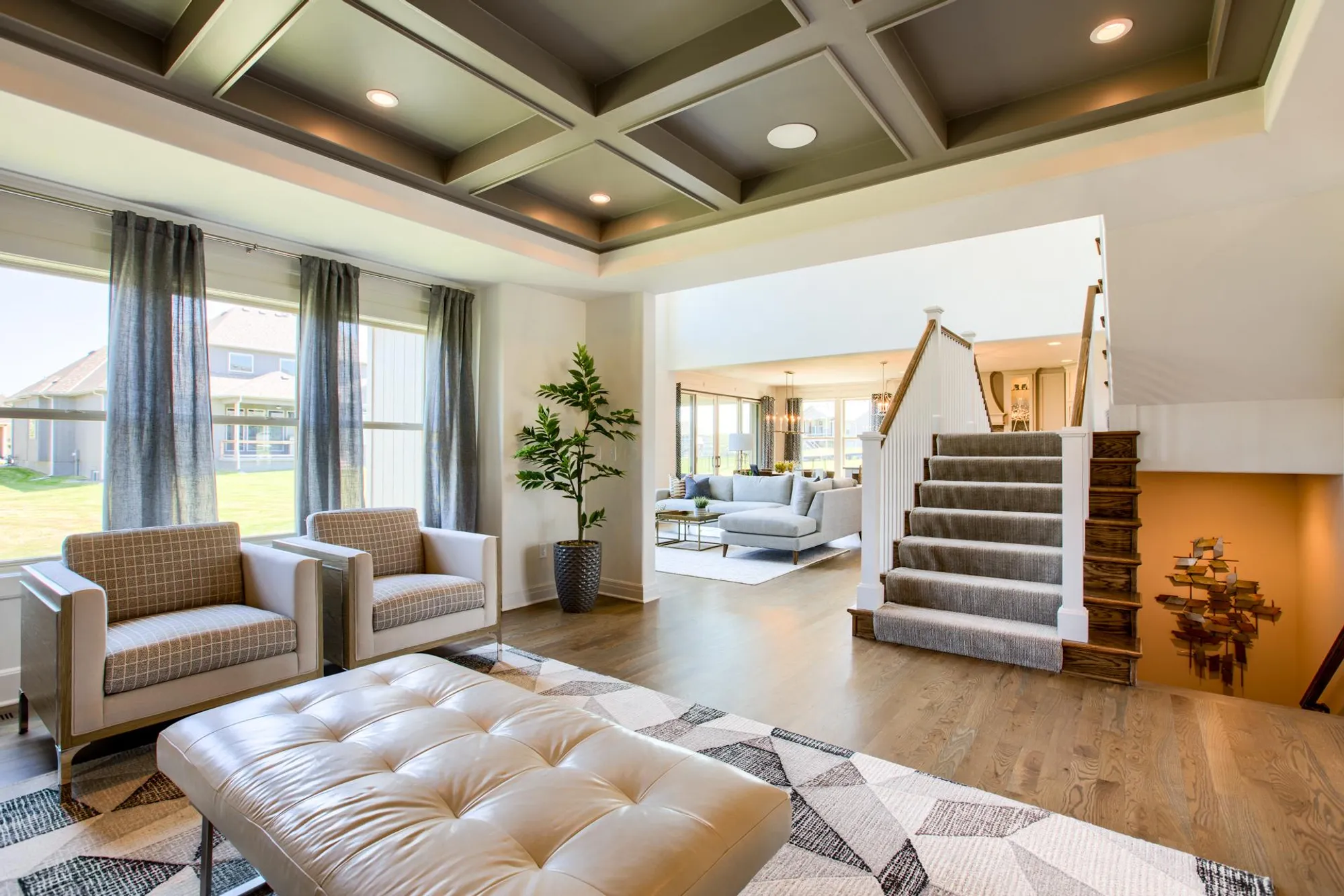Bright open living room with gray upholstered chairs, tufted ottoman, large windows with sheer curtains, and a staircase on the right.