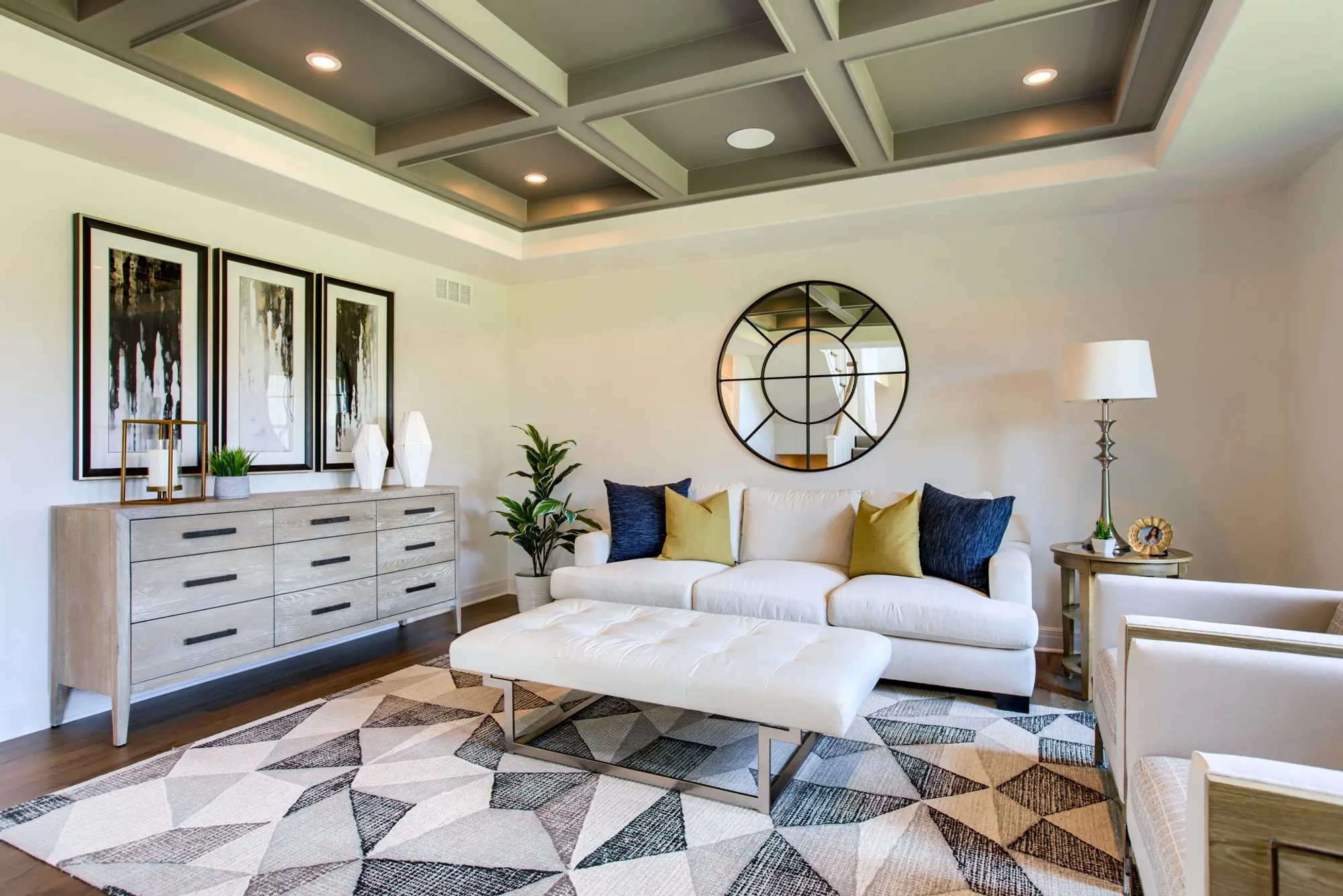 Modern living room with a white sofa, geometric-pattern rug, and round wall mirror above a light wood dresser.