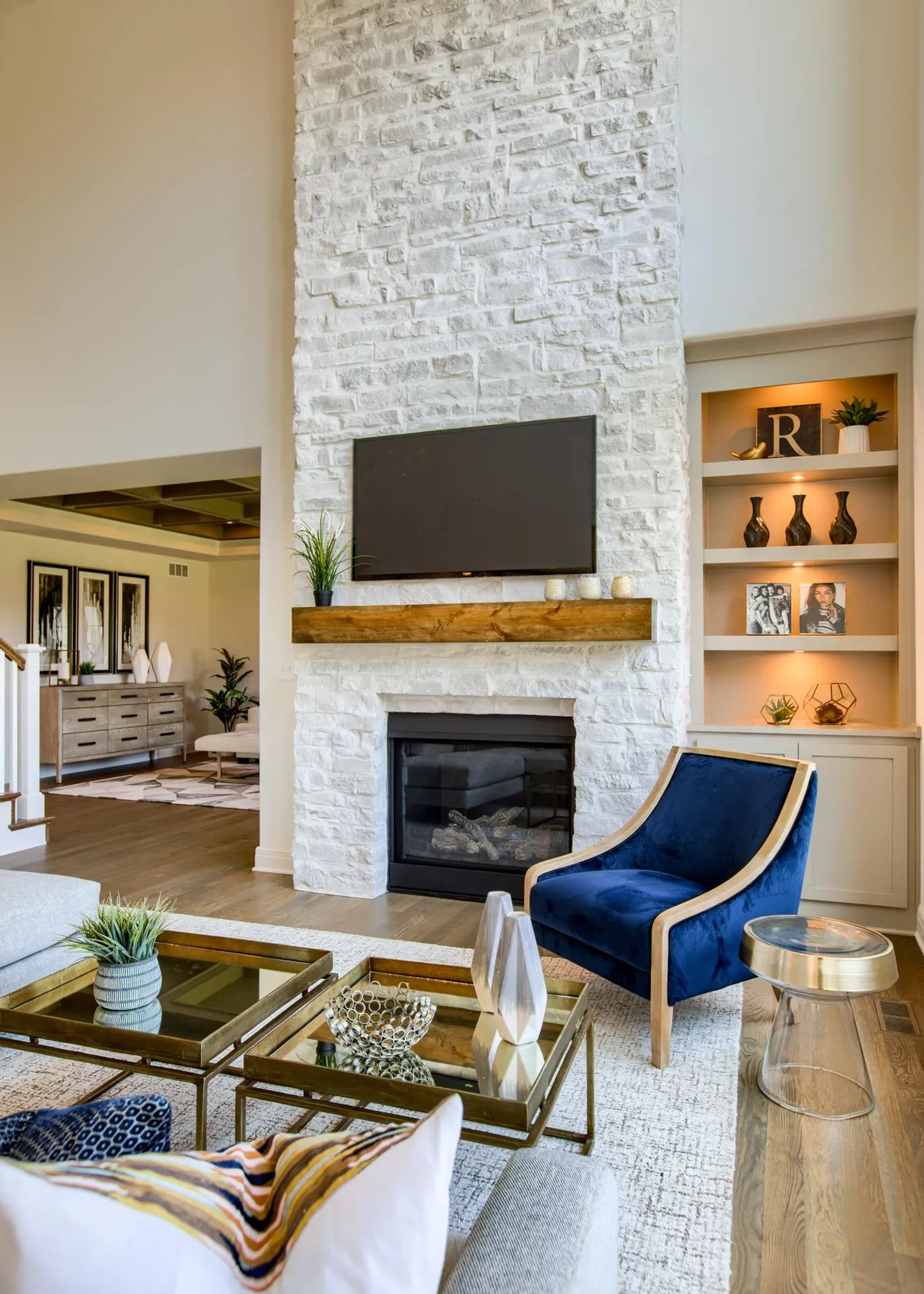 Living room with a white stone fireplace, wall-mounted TV above a wooden mantel, and a blue velvet chair with built-in shelves nearby.