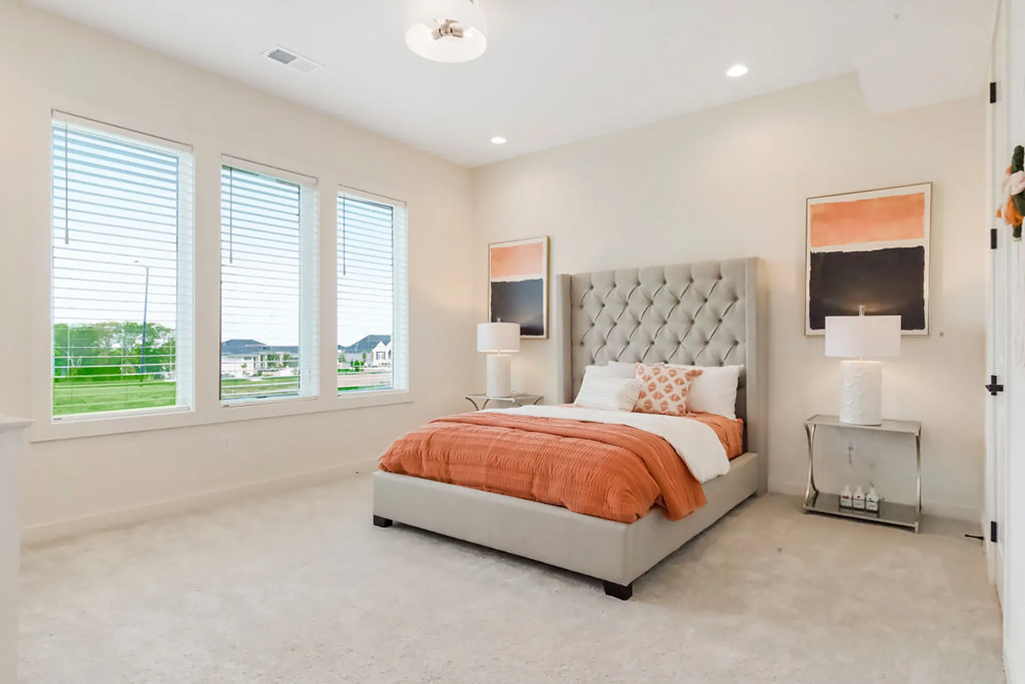 Bright bedroom with a gray tufted bed, orange bedding, two nightstands, and large windows letting in natural light.