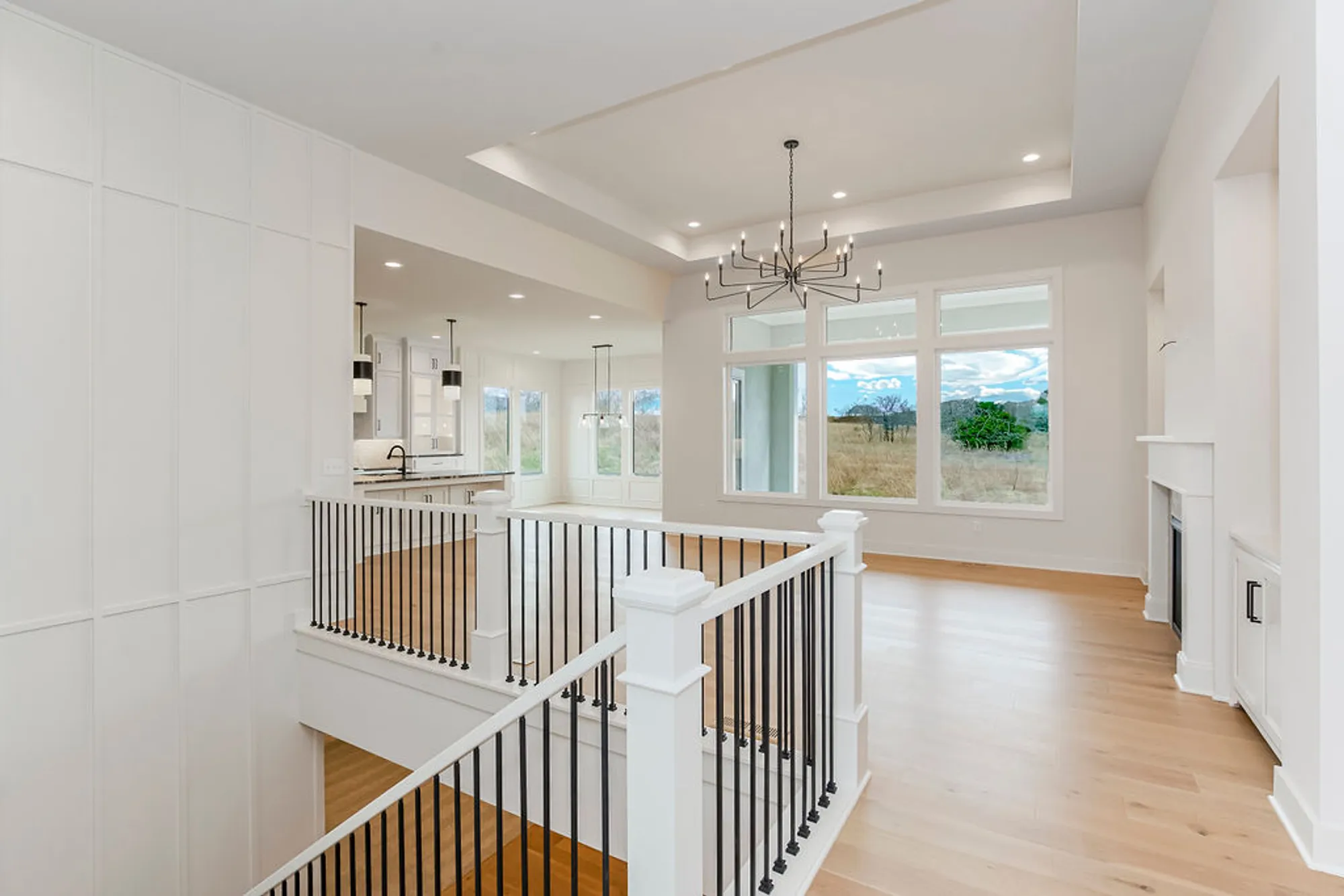 Bright two-level, open living space with white walls, a black metal railing, and a chandelier overlooking a sunny kitchen and large windows to the outdoors in the distance.