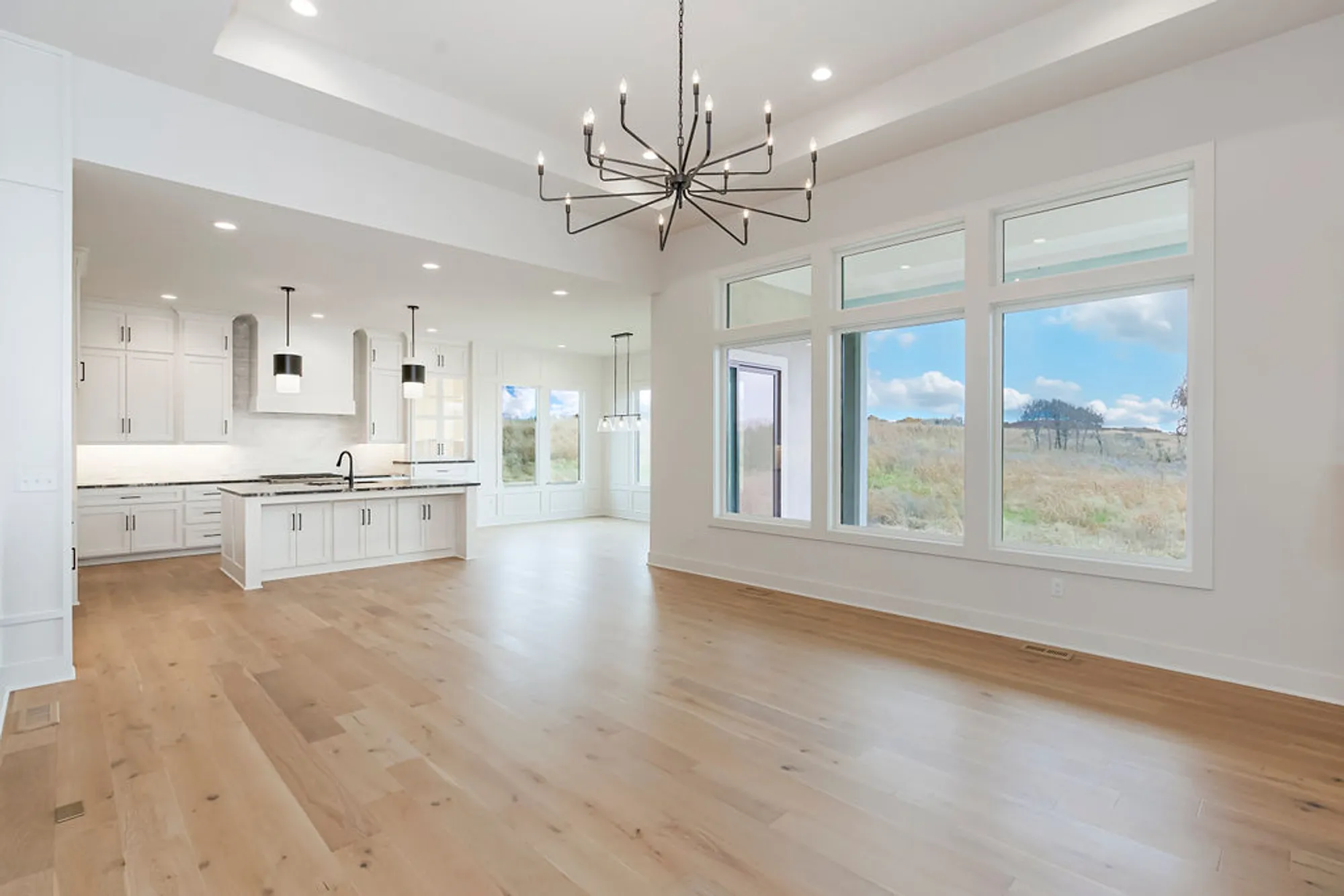 Bright, modern kitchen with white cabinets and a central island beneath a black chandelier, large windows to the outdoors.