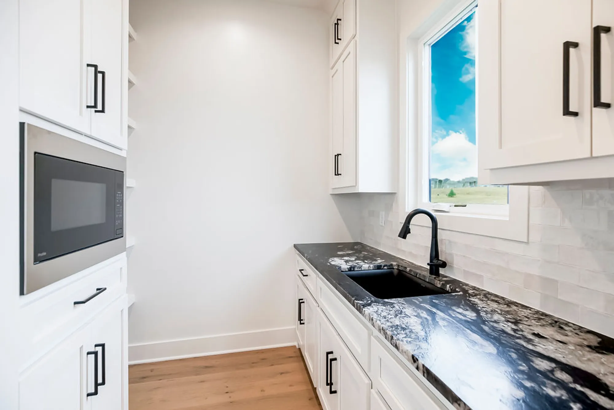 Bright white kitchen with dark marble countertop, black cabinet handles, and a window showing a blue sky and green landscape outside.