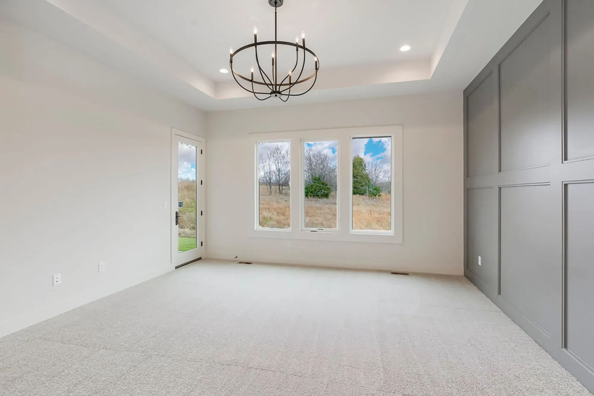 Bright empty room with a circular metal chandelier, three windows, and a door to the outside; soft gray walls and built-in cabinets on the right side.
