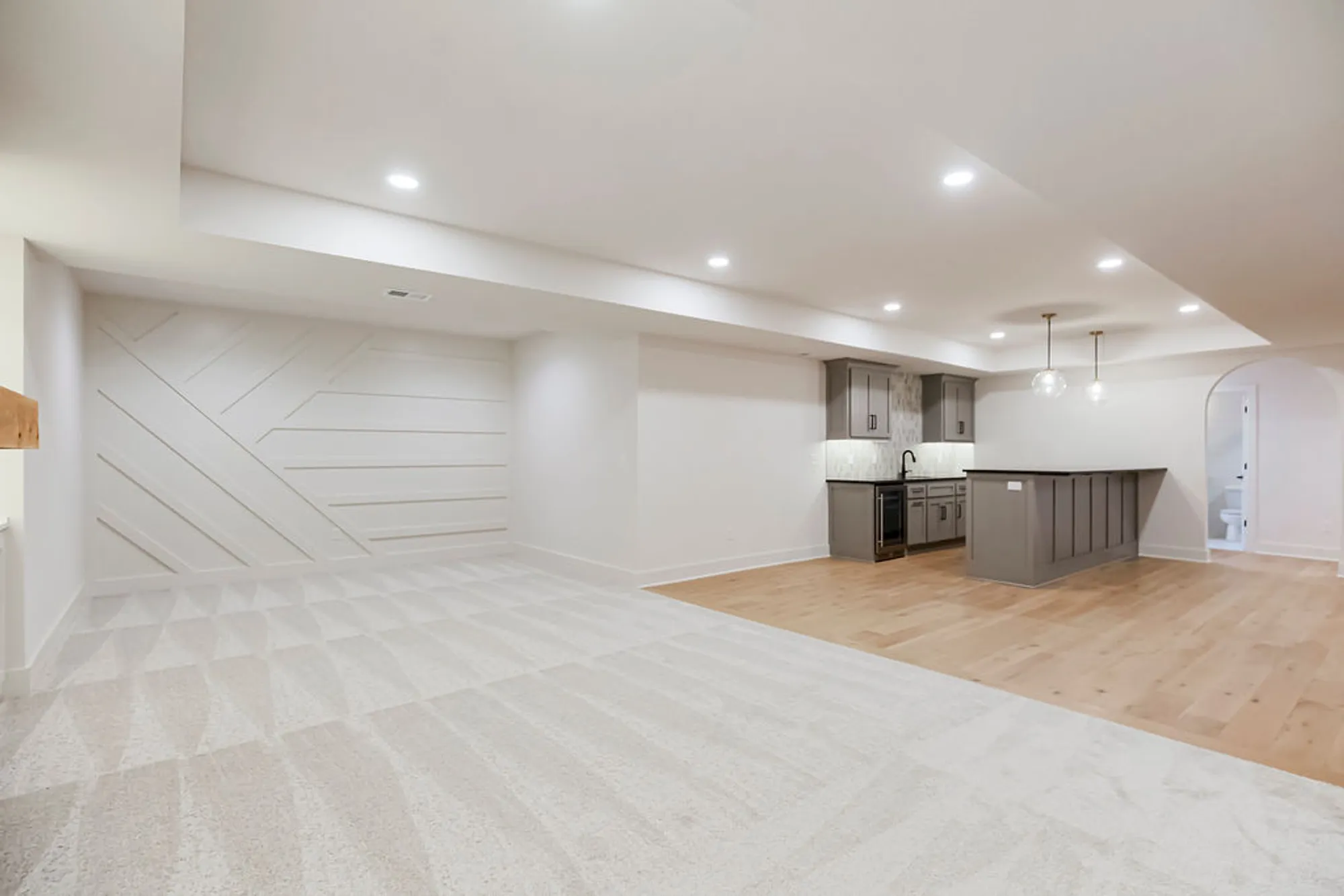 Open-concept kitchen and living area with a gray island, dark cabinets, pendant lights, and mixed carpet and wood flooring in a modern space.