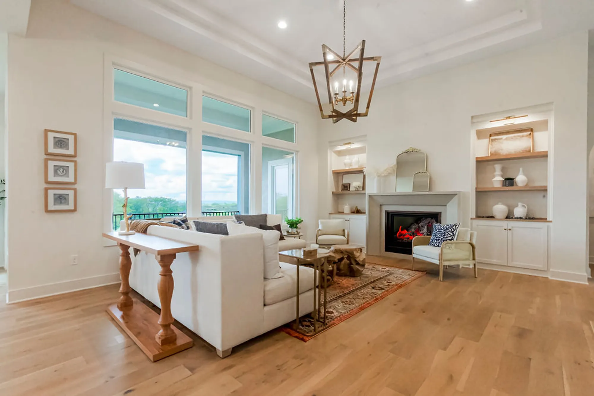 Bright living room with vaulted white ceiling, large windows, and a fireplace flanked by built-in shelves. A beige sofa faces a coffee table and chairs, with natural wood floors and a geometric chandelier above.