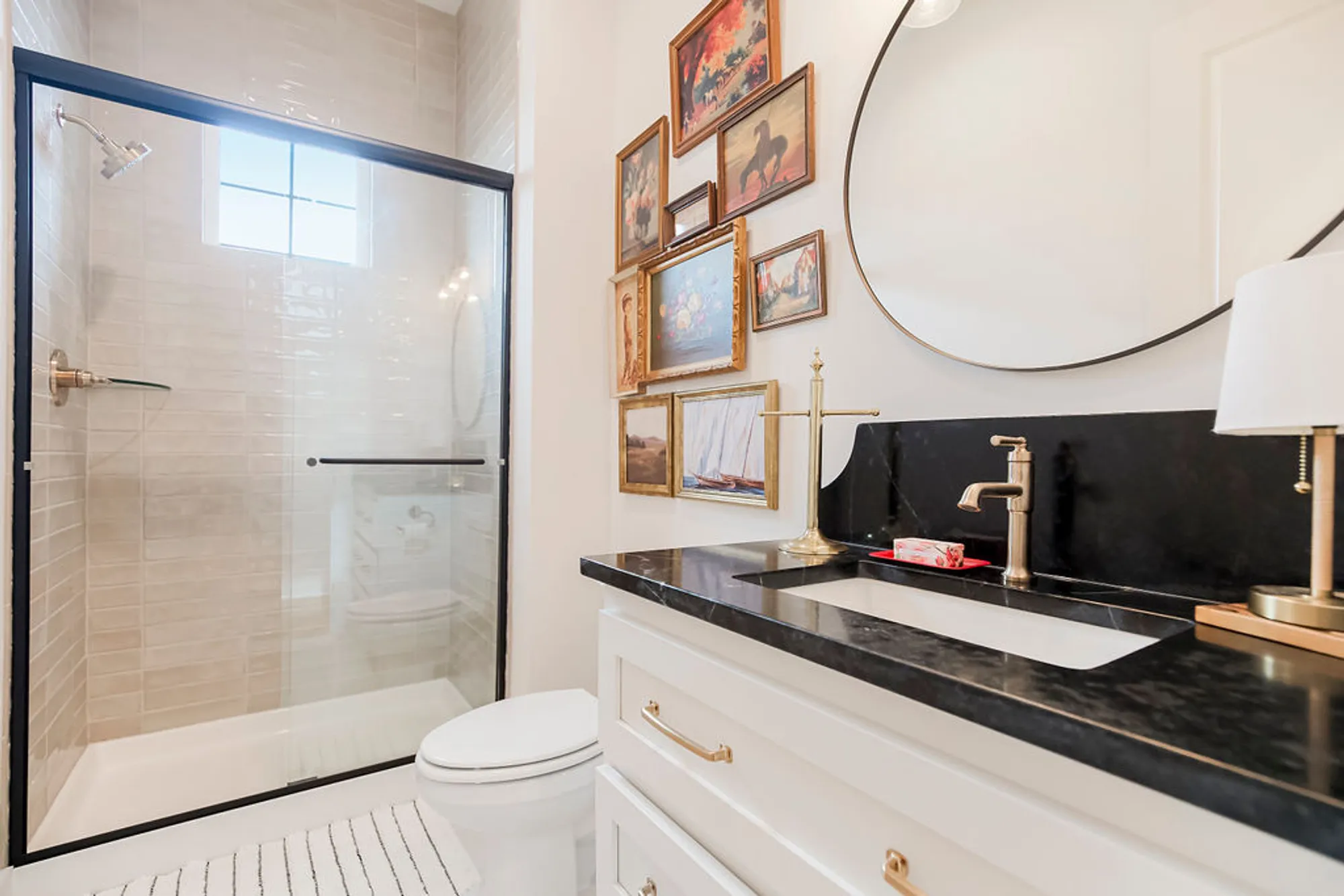 Modern bathroom with glass shower, white subway tiles, and a black countertop vanity with brass hardware; round mirror and a gallery wall of framed art.