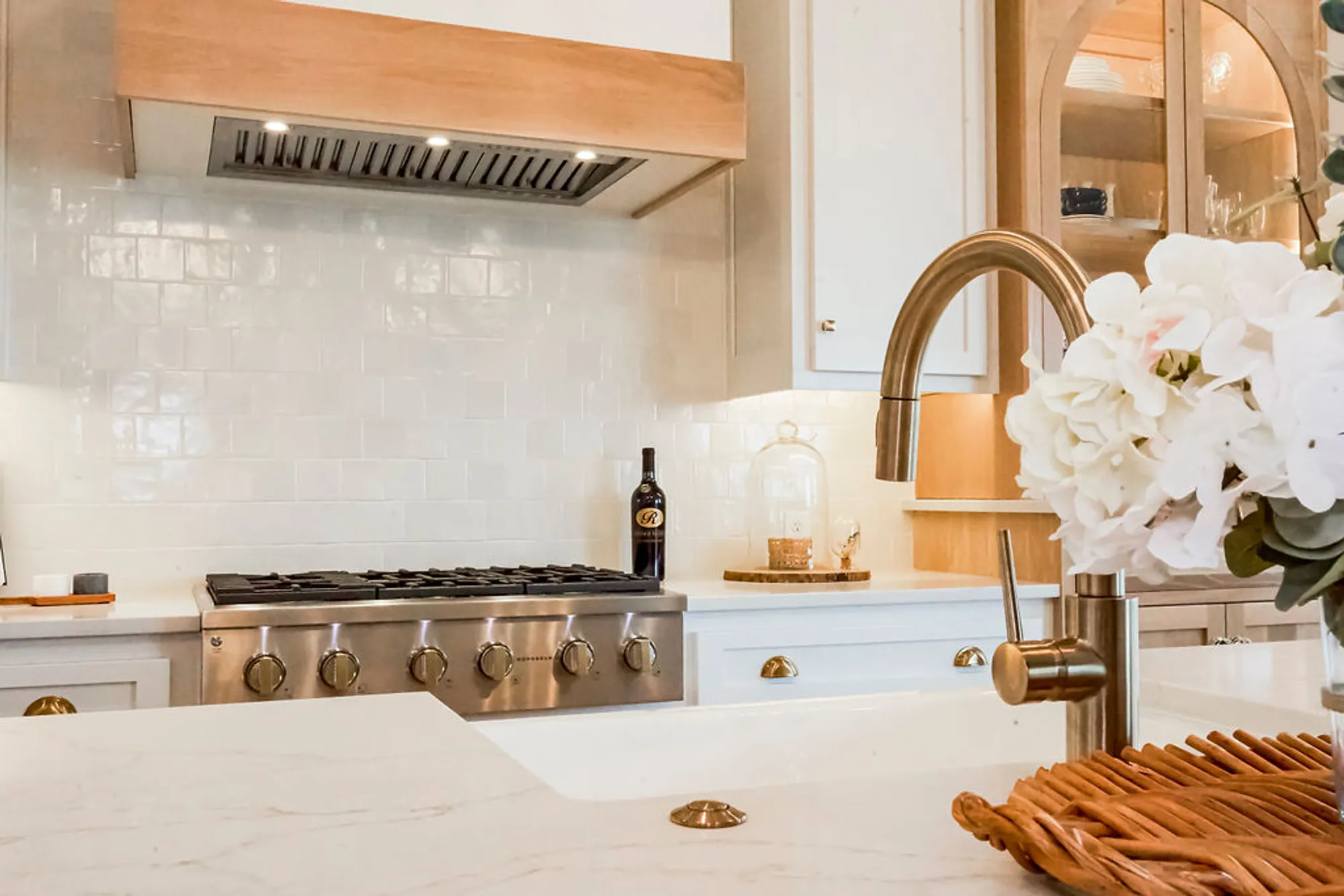 Modern kitchen scene featuring a stainless gas range, white tile backsplash, wooden range hood, and a brass faucet beside a bouquet of white flowers on the counter.