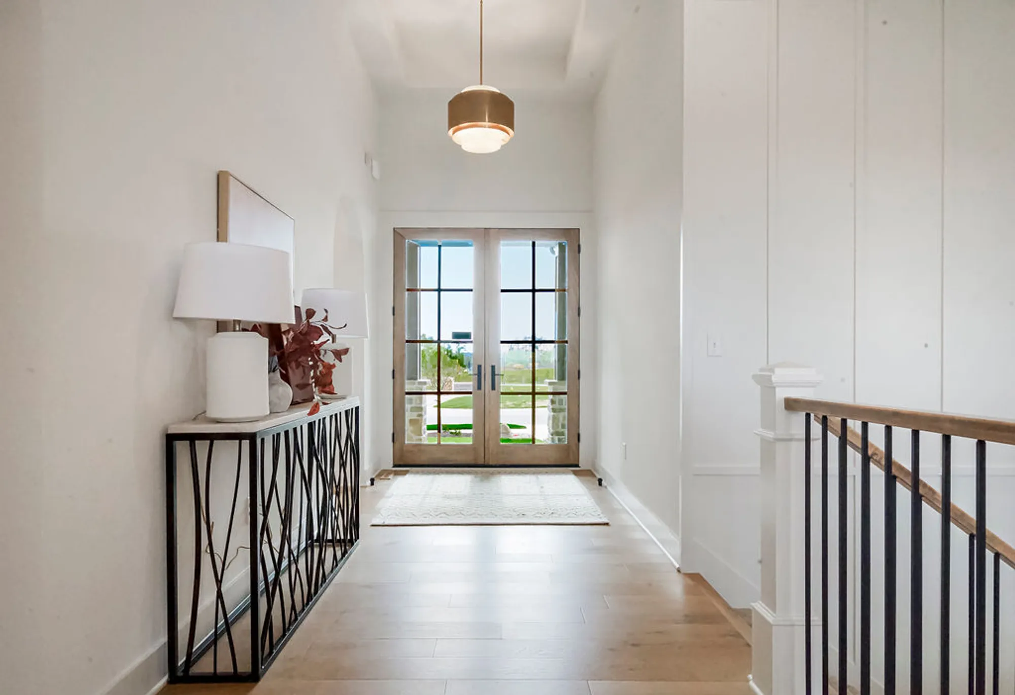 Bright hallway with white walls, a black geometric railing, and glass-paneled double doors at the end, letting in daylight.