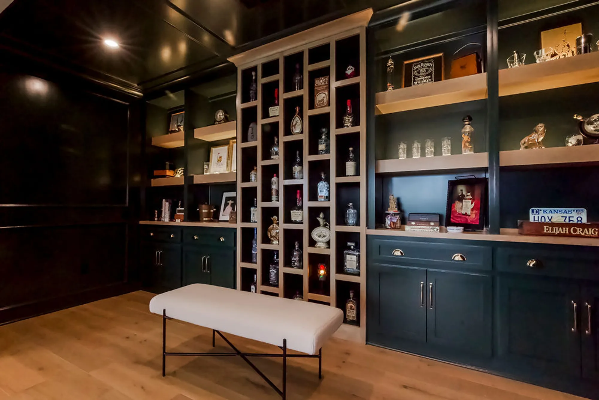 A dark home bar wall with built‑in shelves displaying whiskey bottles and framed items, and a white cushioned bench in the foreground.