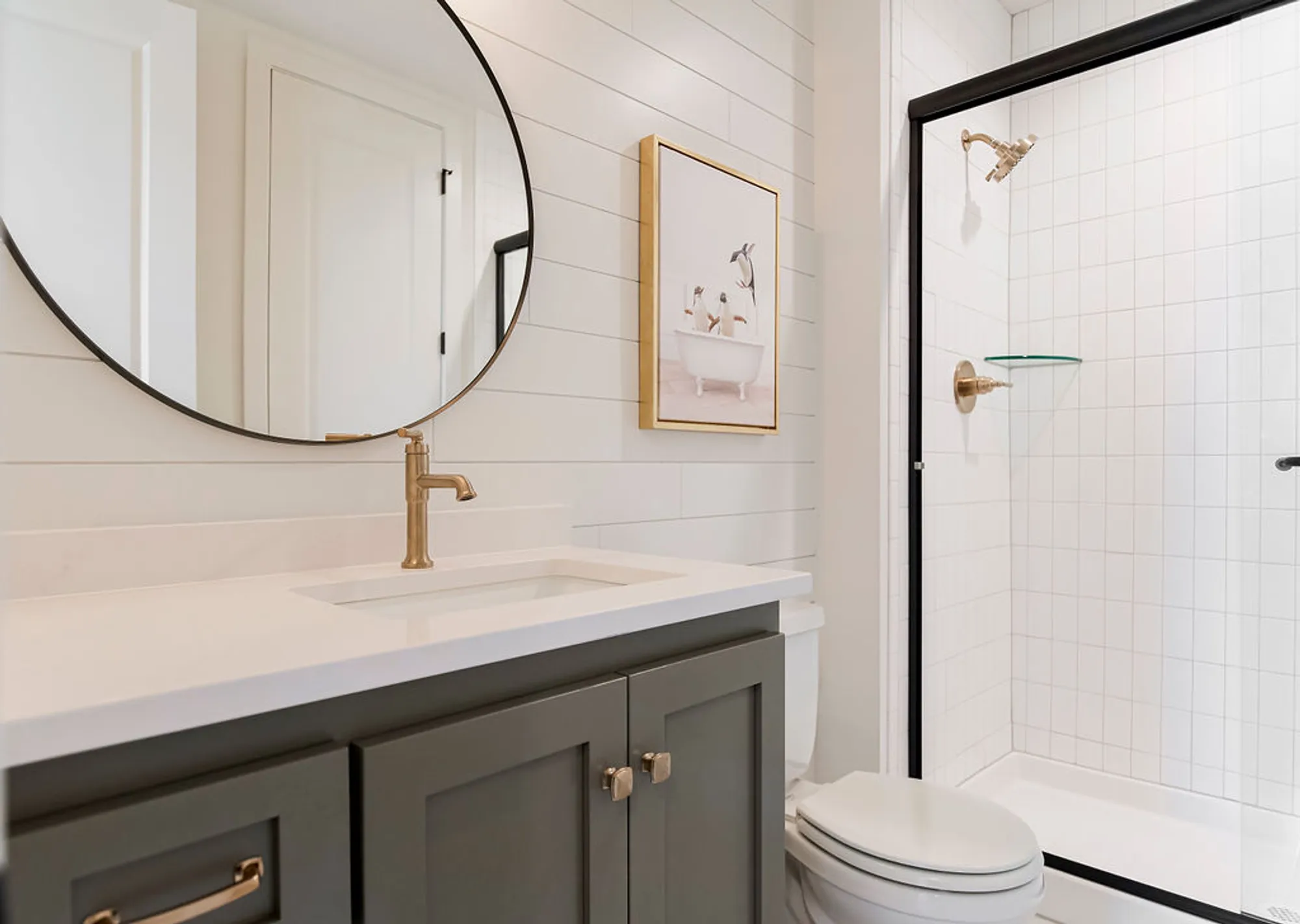 Modern bathroom with a round mirror above a white countertop, gold faucet, dark gray vanity, framed artwork, and a glass shower with a black frame.