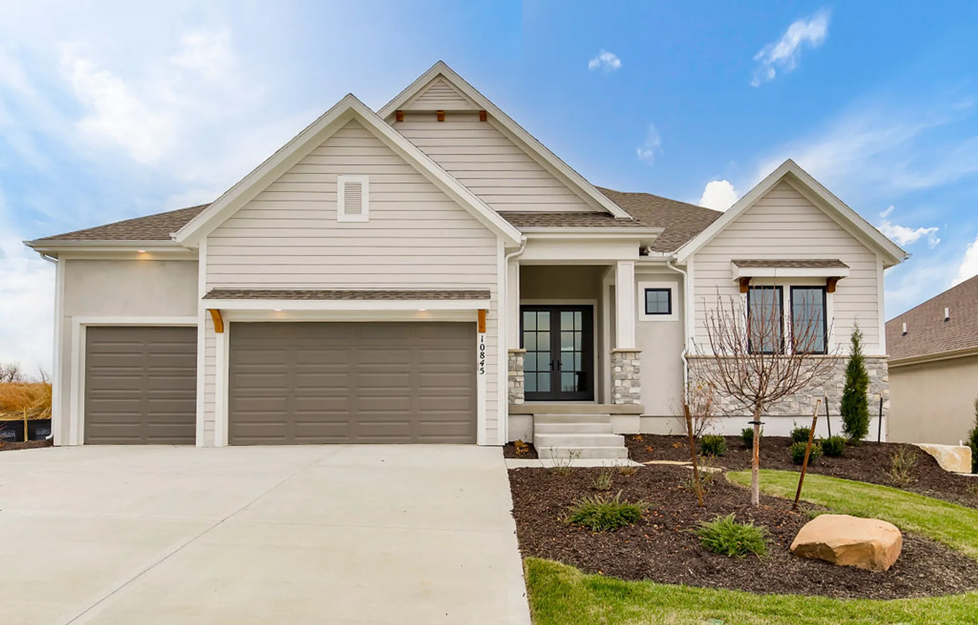 Front view of a modern beige suburban home with a two-car garage and a manicured yard under a blue sky, 10884 address plaque visible by the doorette.