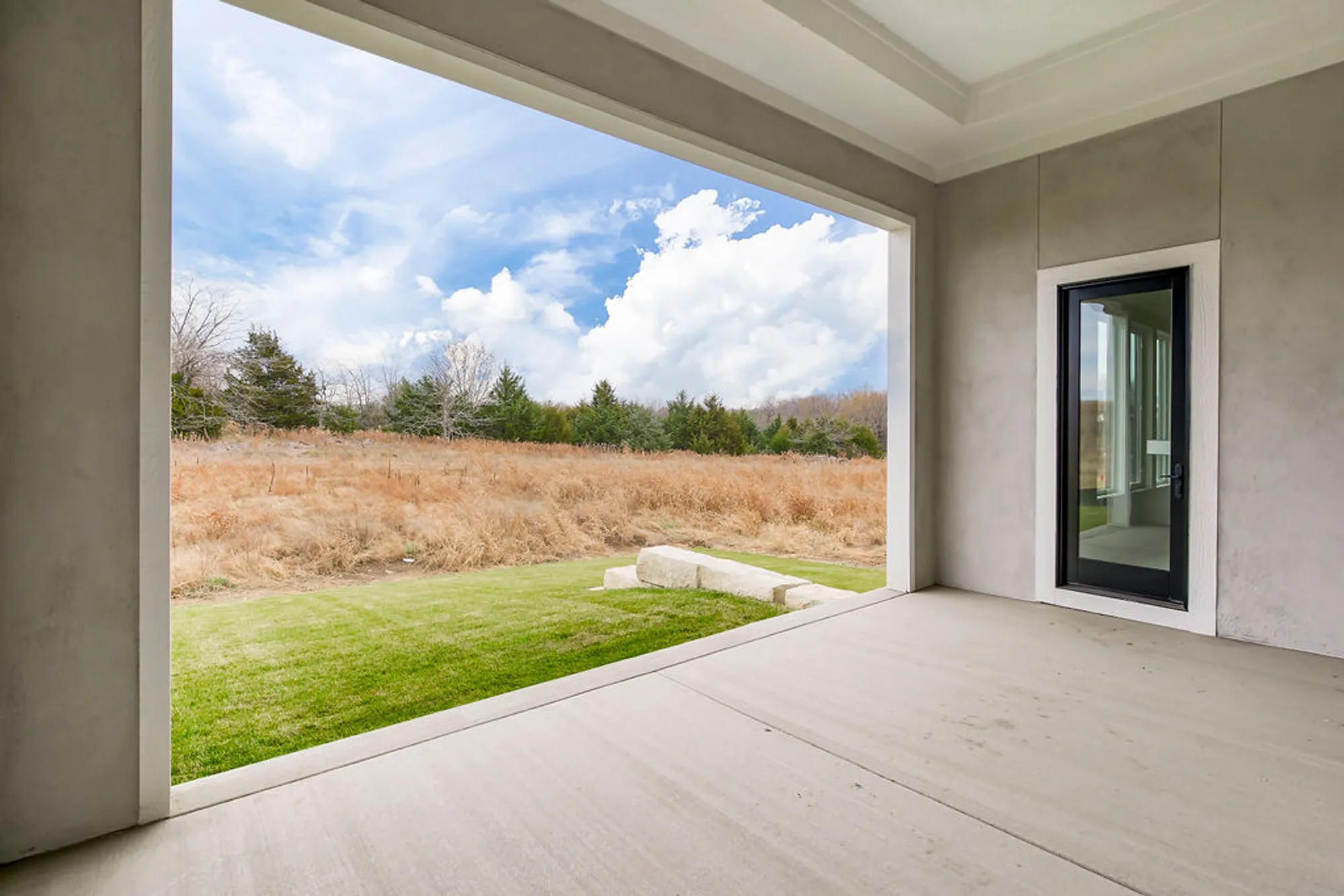 Covered concrete patio with a glass door, open to a grassy yard and fields beyond under a blue sky with clouds.