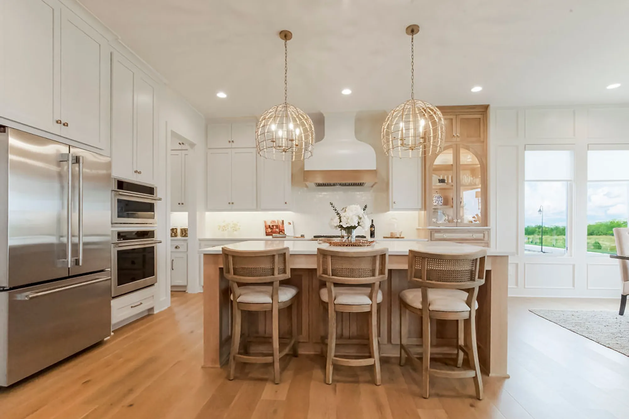 Modern white kitchen with an island, woven pendant lights, and wood stools; stainless steel appliances along the left wall.