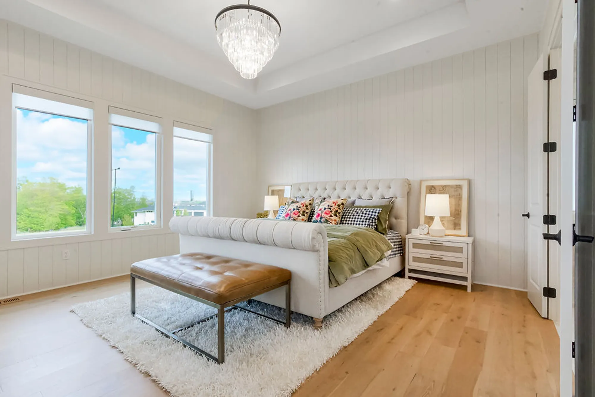 Bright, airy bedroom with a tufted beige bed, decorative pillows, and a crystal chandelier above the bed on white paneled walls and large windows that overlook trees and sky.
