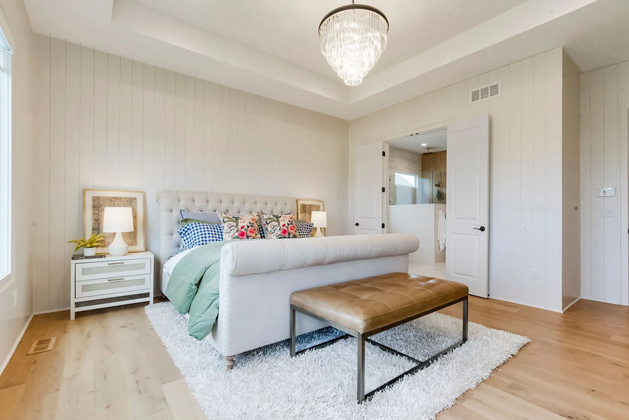 Light-filled bedroom with a tufted upholstered bed, floral and checkered pillows, white nightstands, and a crystal chandelier overhead.