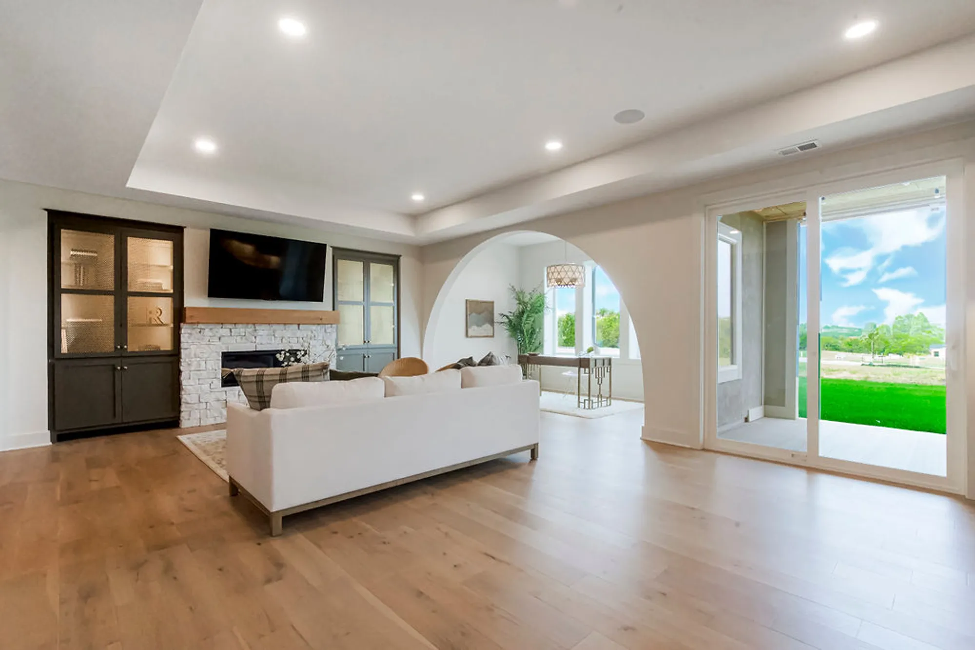 Open-concept living room with white sofa, stone fireplace, dark cabinetry, and large sliding doors to a sunlit backyard.